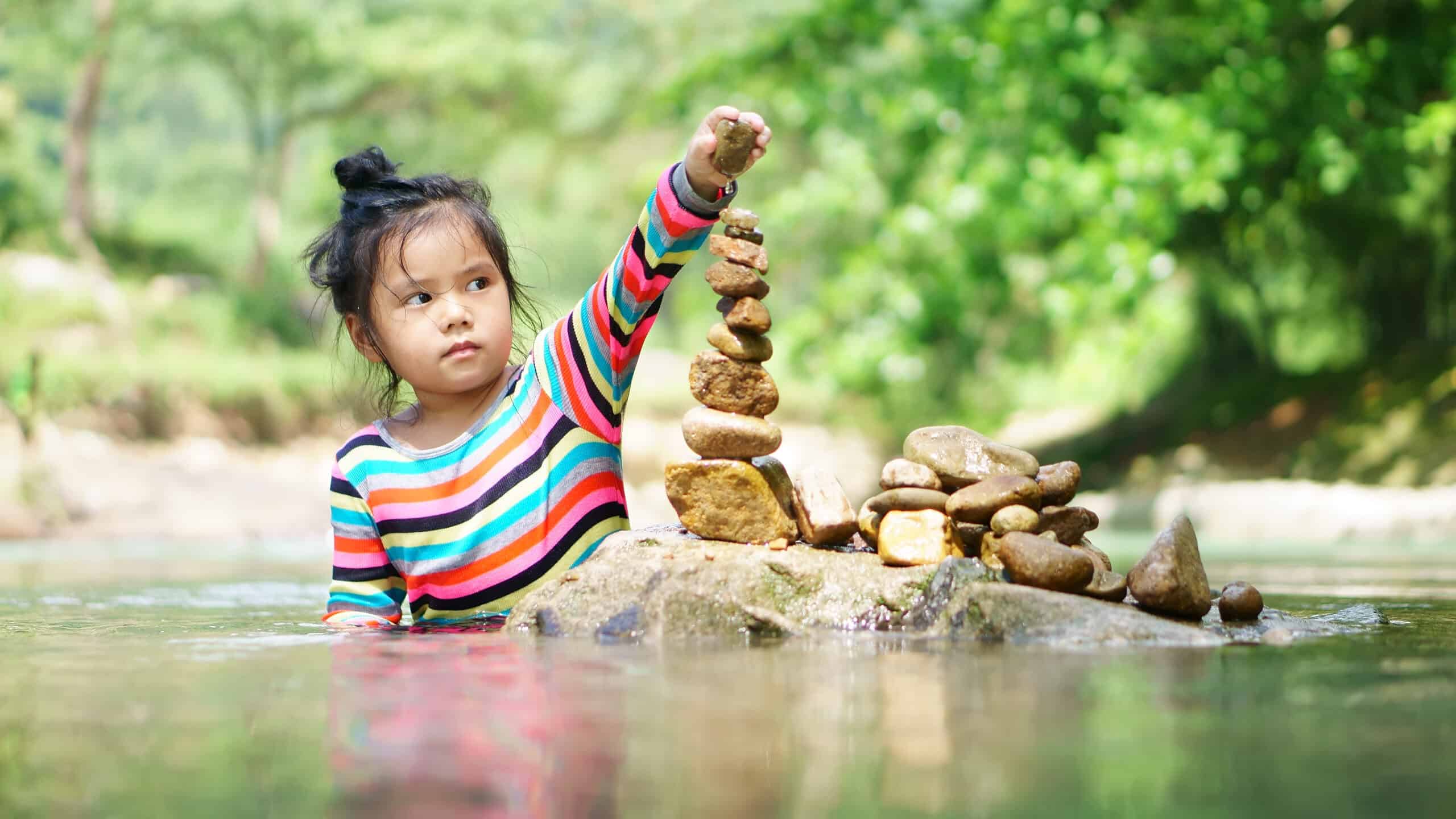 Asian child or kid girl learning sort or stack pebble stone tower or pyramid for water balance training and meditation with nature relax on summer holiday travel at stream or river in forest with tree