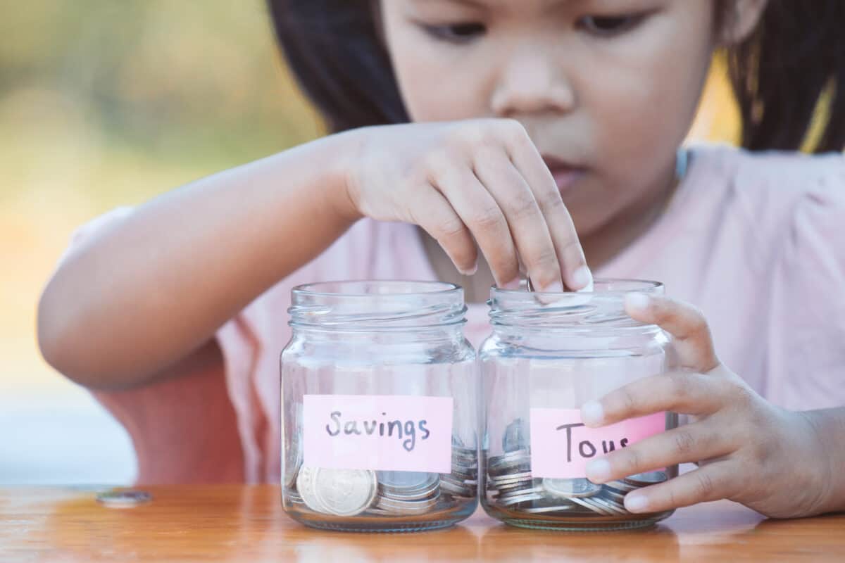 Cute asian little child girl putting coin into glass bottle in the garden. Kid saving money for the future concept