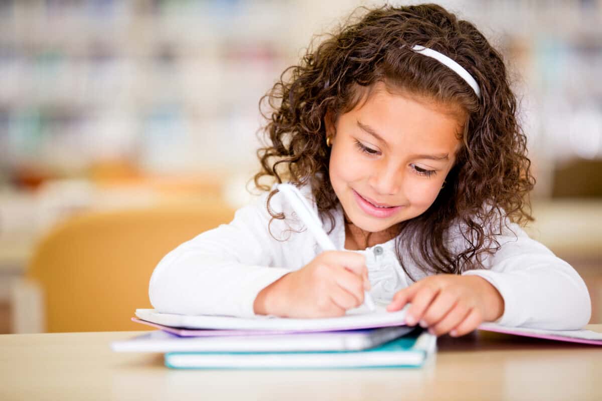 an olive-skinned female presenting child with dark brow curly /wavy should length hair held back with a slender white headband, smiles as she writes something with a white pen or marker in a notebook. The notebook in which she write is in you of two other notebooks one of which is aqua. Indistibct classroom background with chair back visible. 