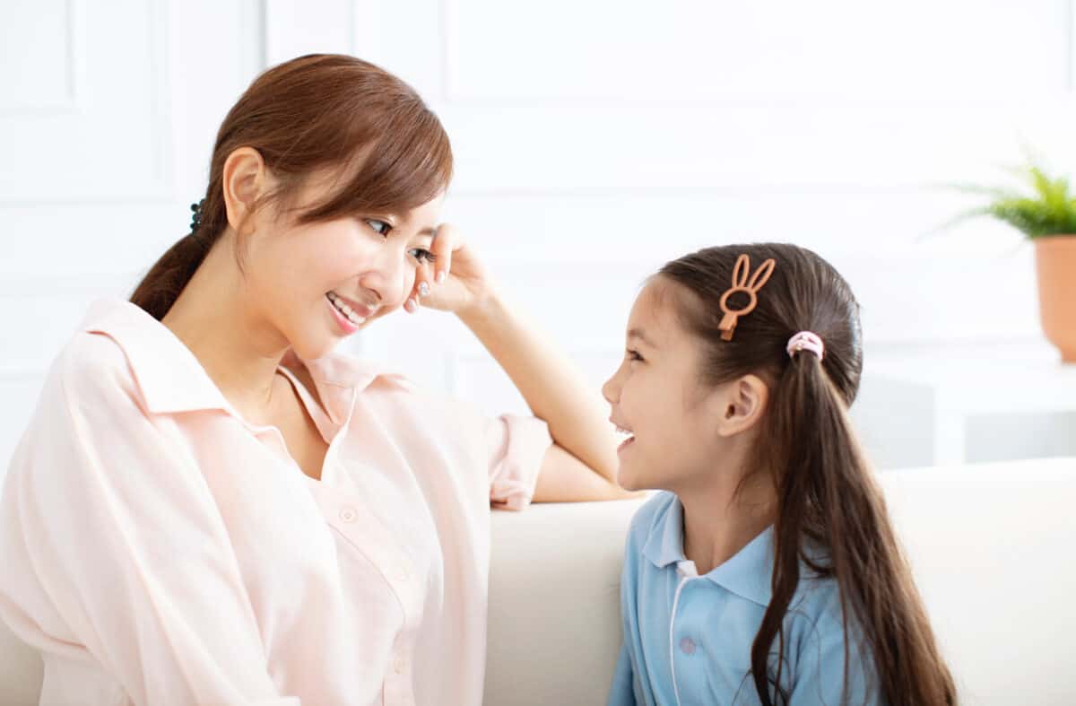 Left frame: A light-skinned Asian woman with long dar hair tied in a ponytail is visible. She is facing center frame. She is wearing a shirt-sleeved pink blouse. Her right arm, in the rear frame, is visible;e propping up her head at her temple. She is smiling. In the right Fram is a little light-skinned Asian girl. She is facing the center frame. Only her Left side is visible. Her hairr is quite long and dark. One long side pony tail is visible. she also has a the outline of a rabbit clip in her hair. She is wearing a light blue polo style shirt. She appears to be talking. Indistince homey background.