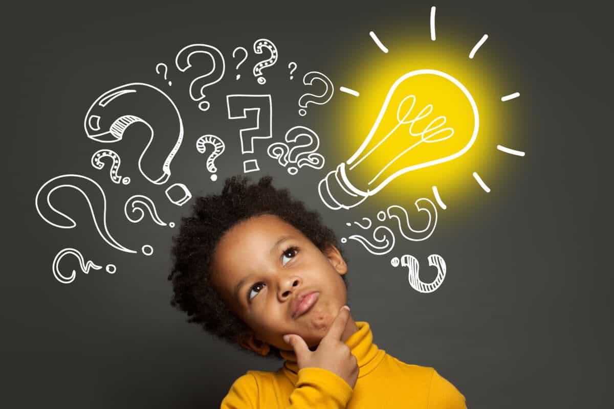 Center bottom frame: A brown-skinned boy with a medium Afro (hairstyle),  dressed in a yellow turtleneck long-sleeved shirt, with his head tilted toward lift frame, has his right hand at his chin with his thumb and index finger extended, his eyes looking up, as if he is thinking . The background is a chalkboard on which question marks of various sizes have been drawn with white chalk, and a lightbulb which is outlined in white but has yellow chalk making it appear to be lighted.