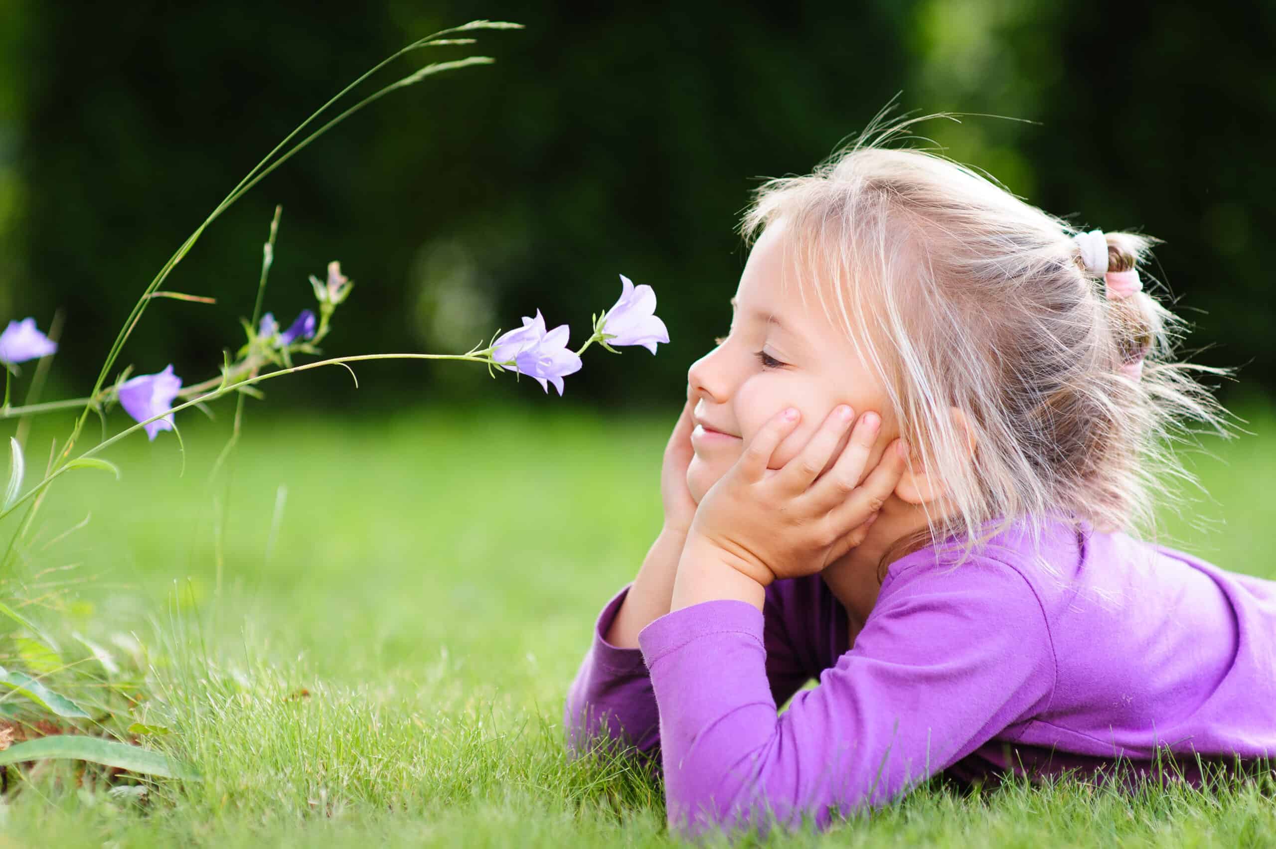 A portrait of a cute little girl lying on the ground in the garden on a nice summer day and looking at a wild flower