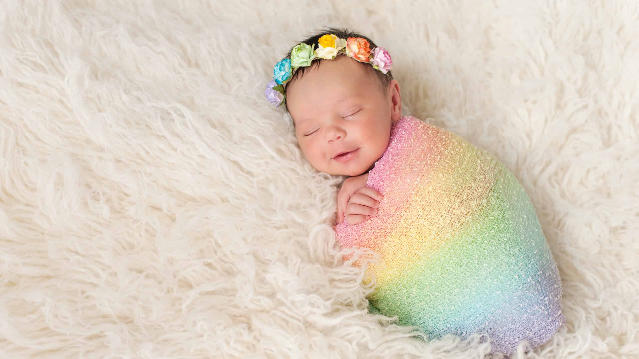 A smiling nine day old newborn baby girl bundled up in a rainbow colored swaddle. She is lying on a cream colored flokati (sheepskin) rug and wearing a crown made of roses.