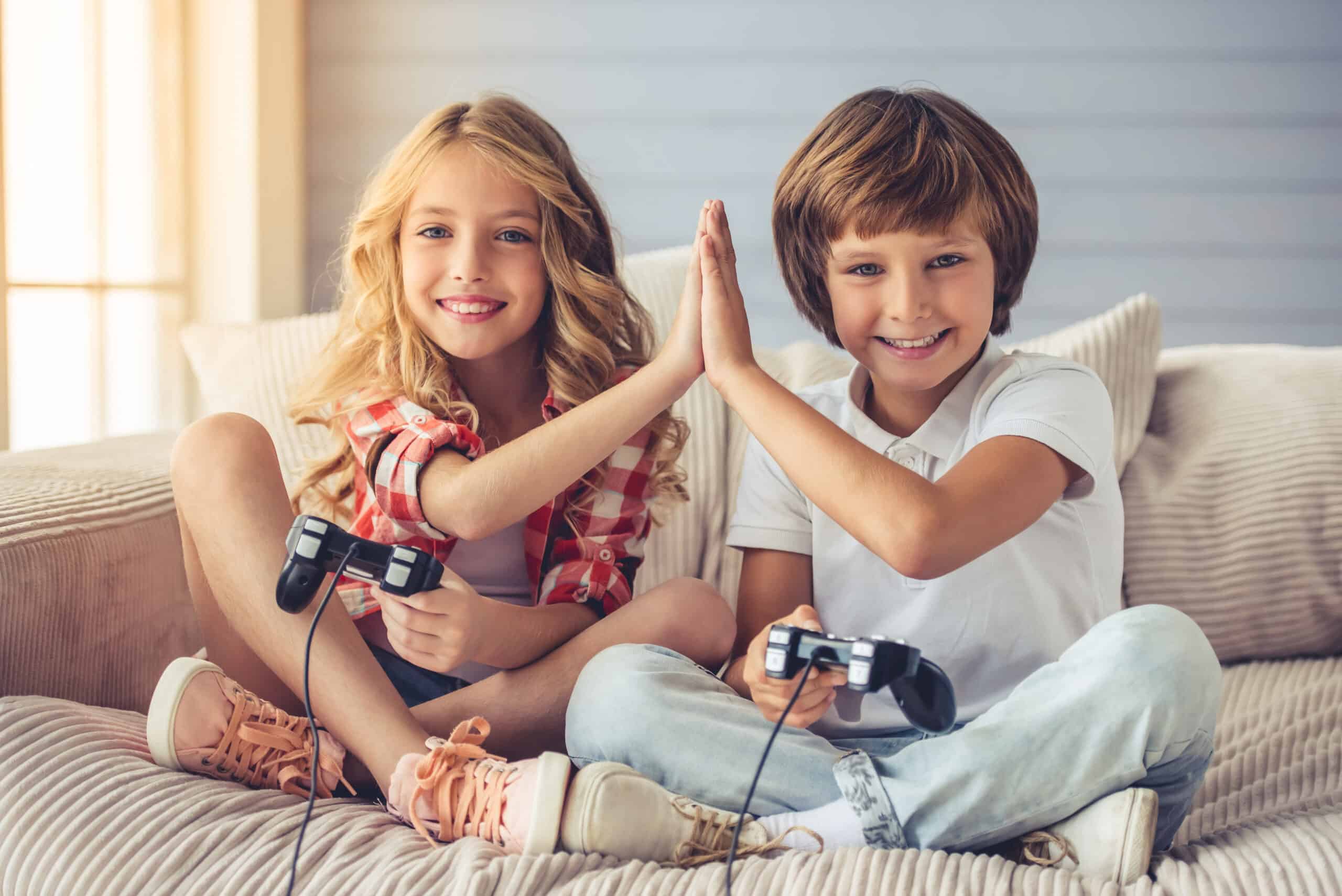 Pretty little girl and boy are playing game console, giving high five, looking at camera and smiling while sitting on sofa at home