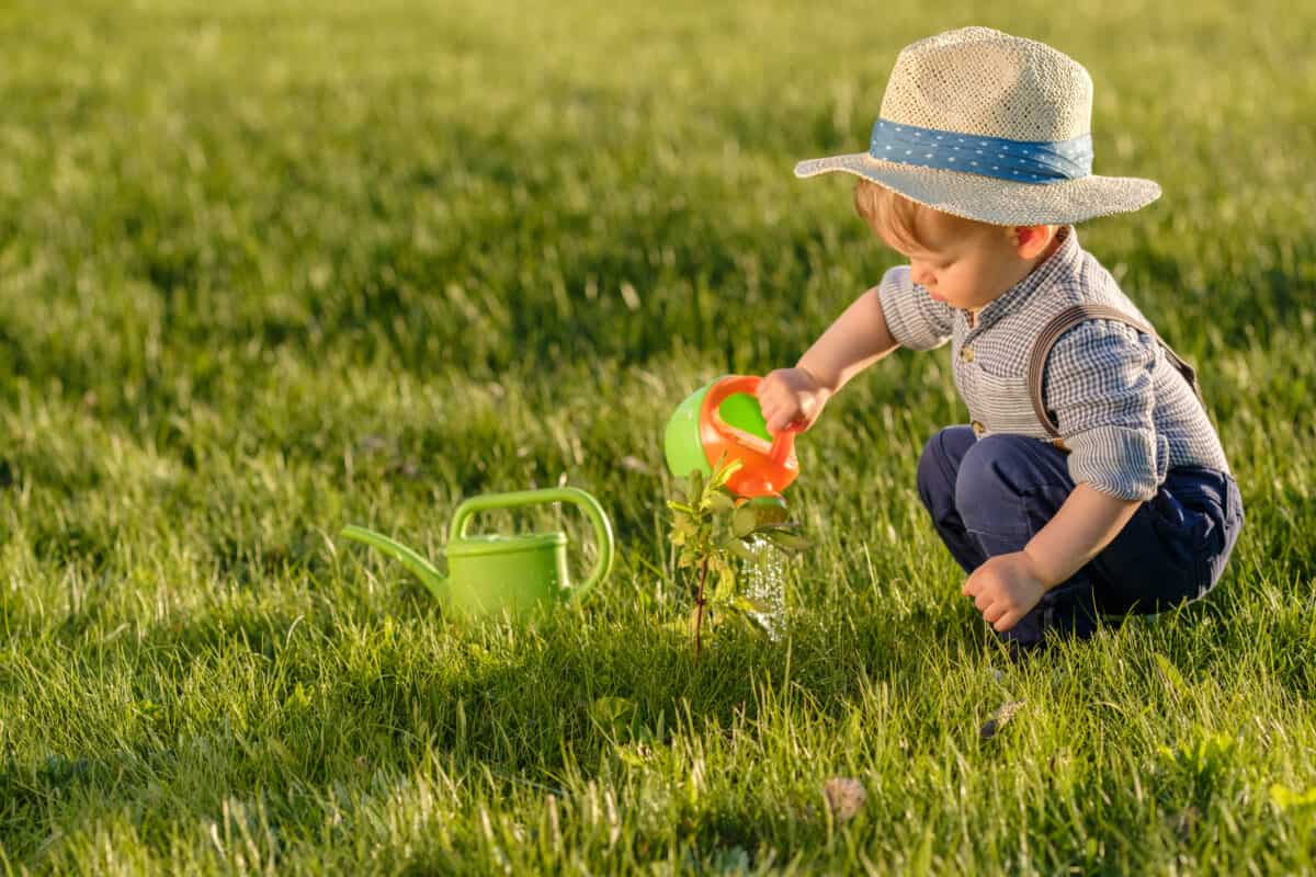 Portrait of toddler child outdoors. Rural scene with one year old baby boy wearing straw hat using watering can