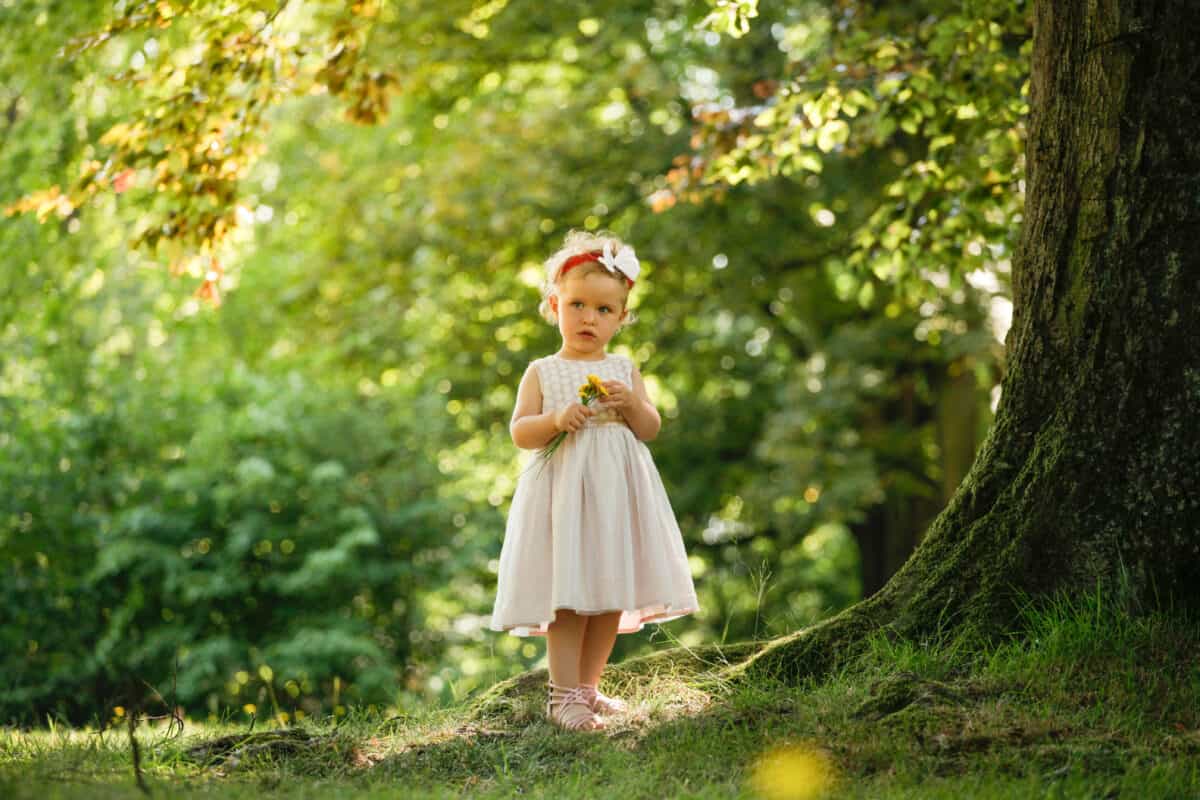Cute little girl standing next to tree in fairy tale forest holding little yellow flower.