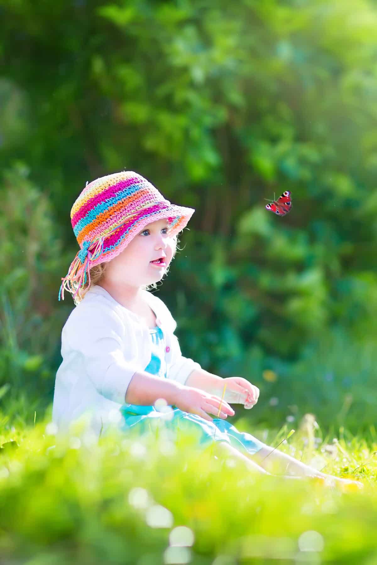 Happy laughing little girl wearing a blue dress and colorful straw hat playing with a flying butterfly having fun in the garden on a sunny summer day