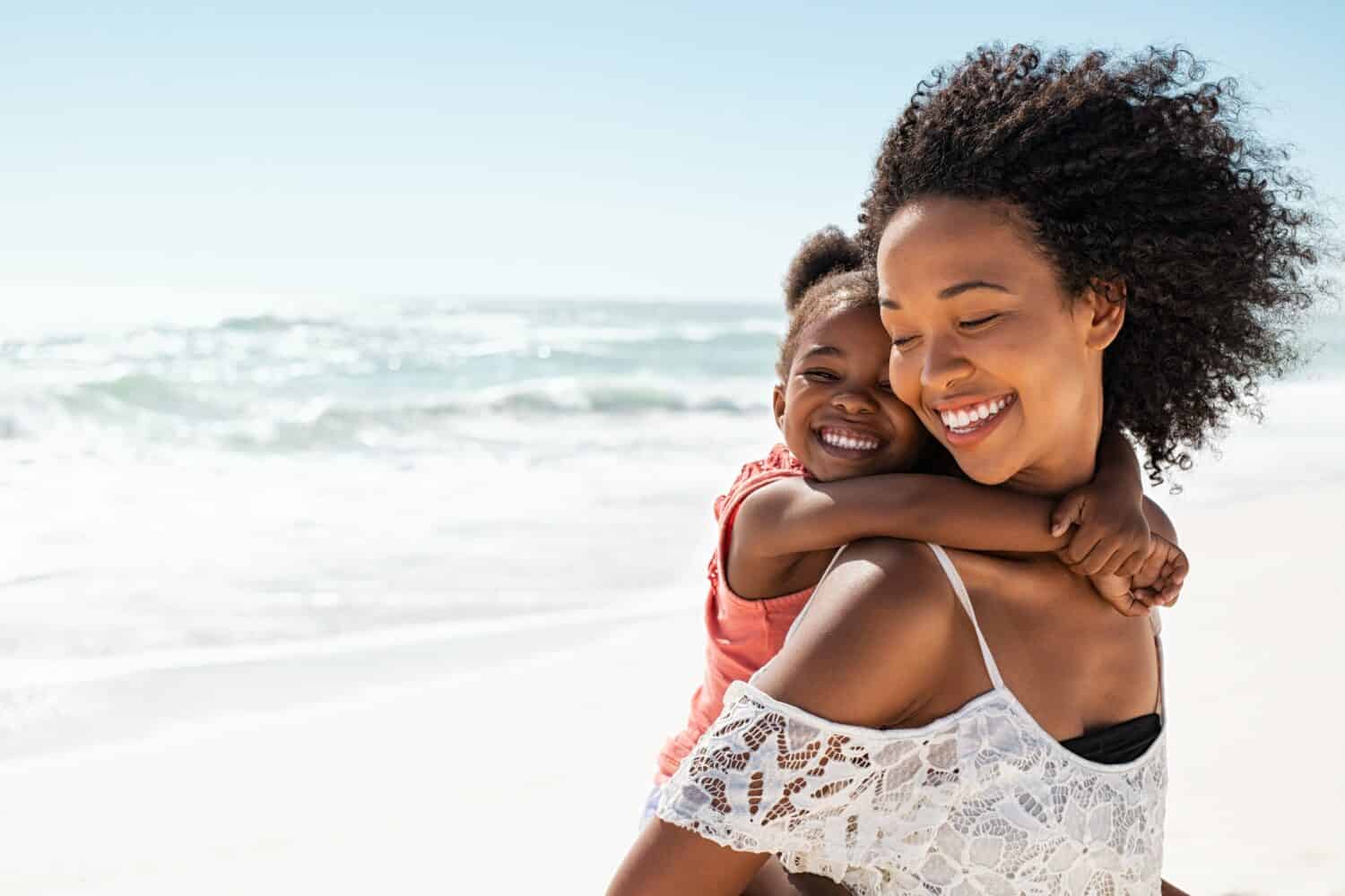 Smiling young black mother and beautiful daughter having fun on the beach with copy space. Portrait of happy sister giving a piggyback ride to cute little girl at seaside. Lovely kid embracing her mom