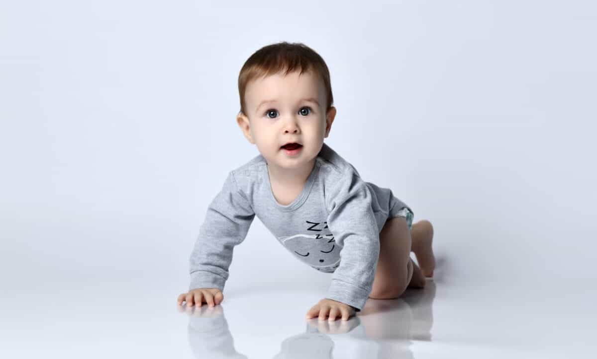 Little baby boy toddler in grey casual jumpsuit and barefoot crawling on floor, smiling and looking up over white wall background. Trendy baby clothing, first steps and happy childhood concept