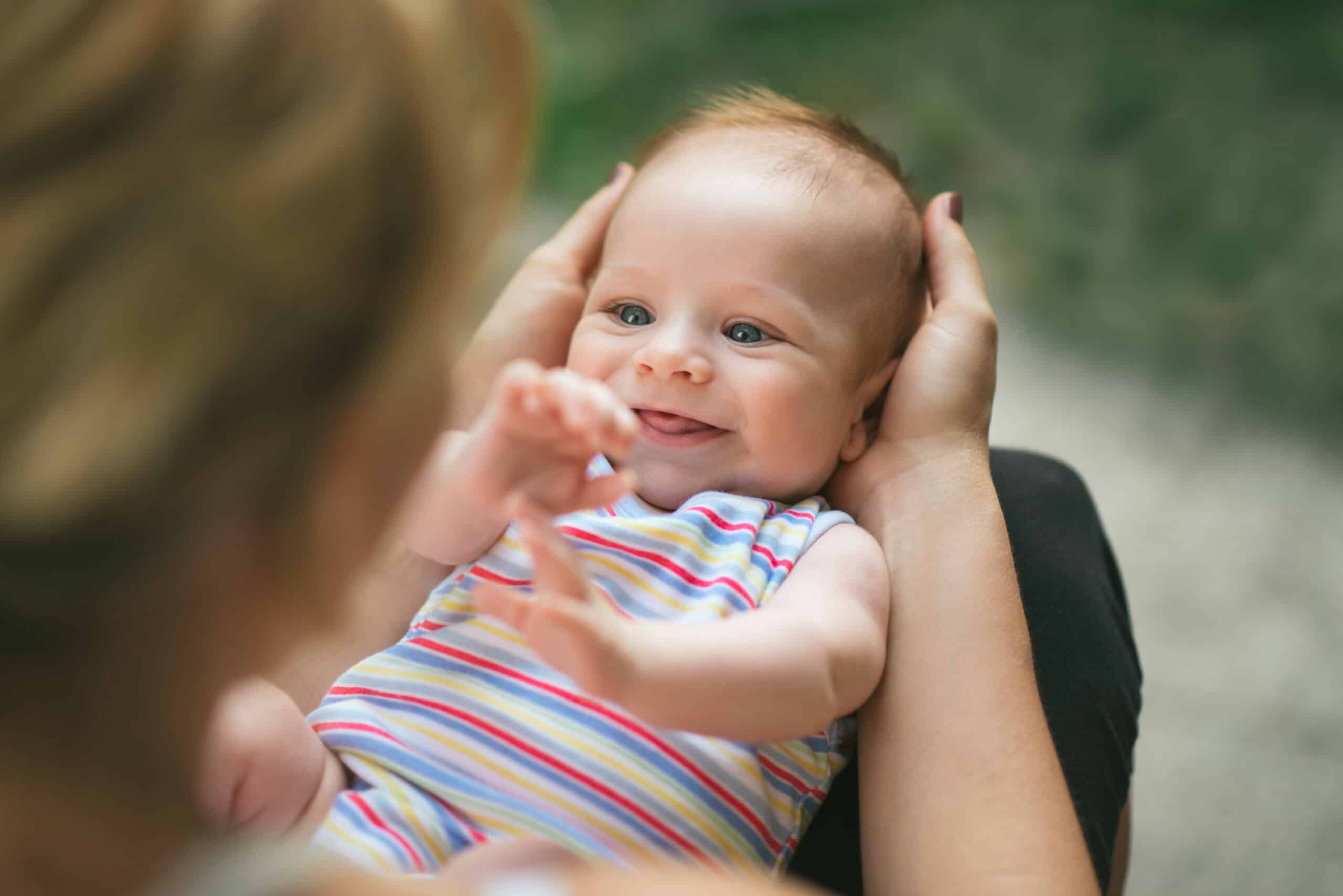 Portrait of a beautiful, laughing baby boy with gorgeous blue eyes being held by his mother, on his back, in her lap in the garden