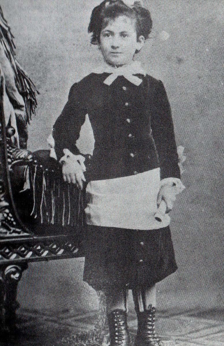 Black and white photo circa 1880 of Maria Montessori (1870-1952). She is visible center frame, sanding next to a chair with her right arm resting on the left arm of the chair. The chair is intricately carved wood, draped in ornate fabric. Maria, who is about 10 yers old, is dressed in a long-sleeved dark(black?) dress that falls below her knees. There is a white collar or scarf tied at her neck. A white apron or similar, is tied around her hips. She is holding a rolled up piece of paper. possibly a diploma or an award.
