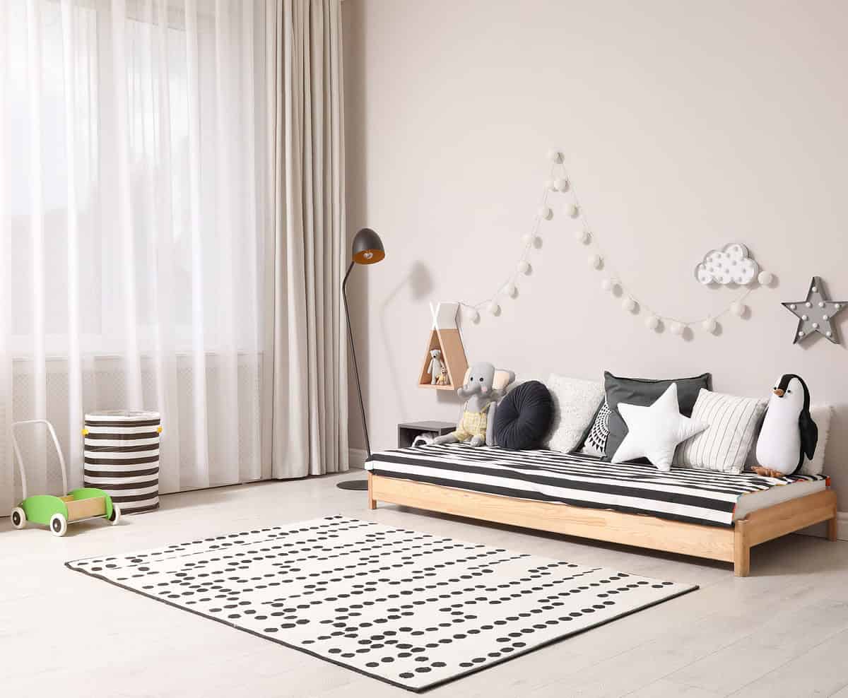 Full frame photograph of a child’s bedroom. At the back of the left frame is a Little wooden pushcart with green sides. Next to the pushcart is a black and white striped laundry hamper against a large window with a sheer curtain of white and a neutral colored blackout curtain. In front of the curtain in center frame back is a lamp. On the right side of the frame against a very lightly color taupe wall is a low bed with seven pillows, including a white star shaped one. The pillows are all neutral colors. A stuffed penguin and a stuffed elephant are visible among the pillows. The bedspread is black and white stripe. The bed itself is made of blonde wood. In front of the bed is a mat or rug that is white with black to grey circles in various patterns the floor is light. Behind the bed hanging on the wall is a wooden triangle shape with a doll in it some white circles that may be lights in a cloud and a star that appeared to have some lights in them