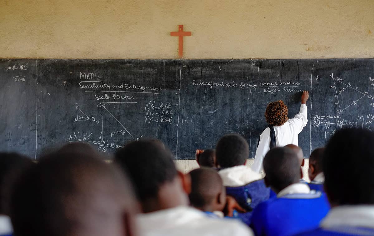 A traditional classroom setting, full frame -  the backs of nine students head are visible. The children are dark-skinned. A teacher is at the front of the classroom, which is in the back of the frame. Her back is turned to th camera nd she is writing on a black chalkboard. there is a blonde/orange wooden cross center ed above the chalkboard. 