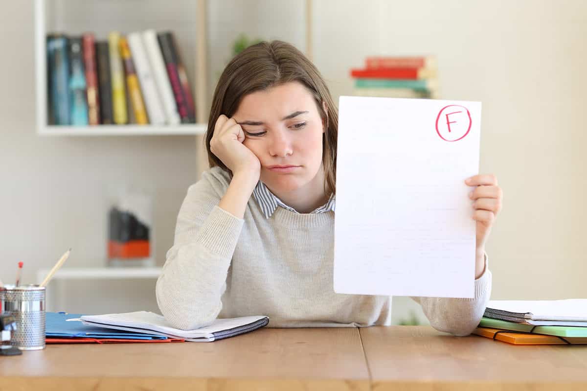 Center frame: light-skinned girl with long straight brown hair is visible seated at a desk with her elbows propped up on the desk; her right hand is propping up her head, while in the right frame her left hand is holding up what appears to be a blank piece of paper with the red letter F in the top right hand corner in a red circle. The girl is not happy. Some out of focus books on a shelf are seen in the background.