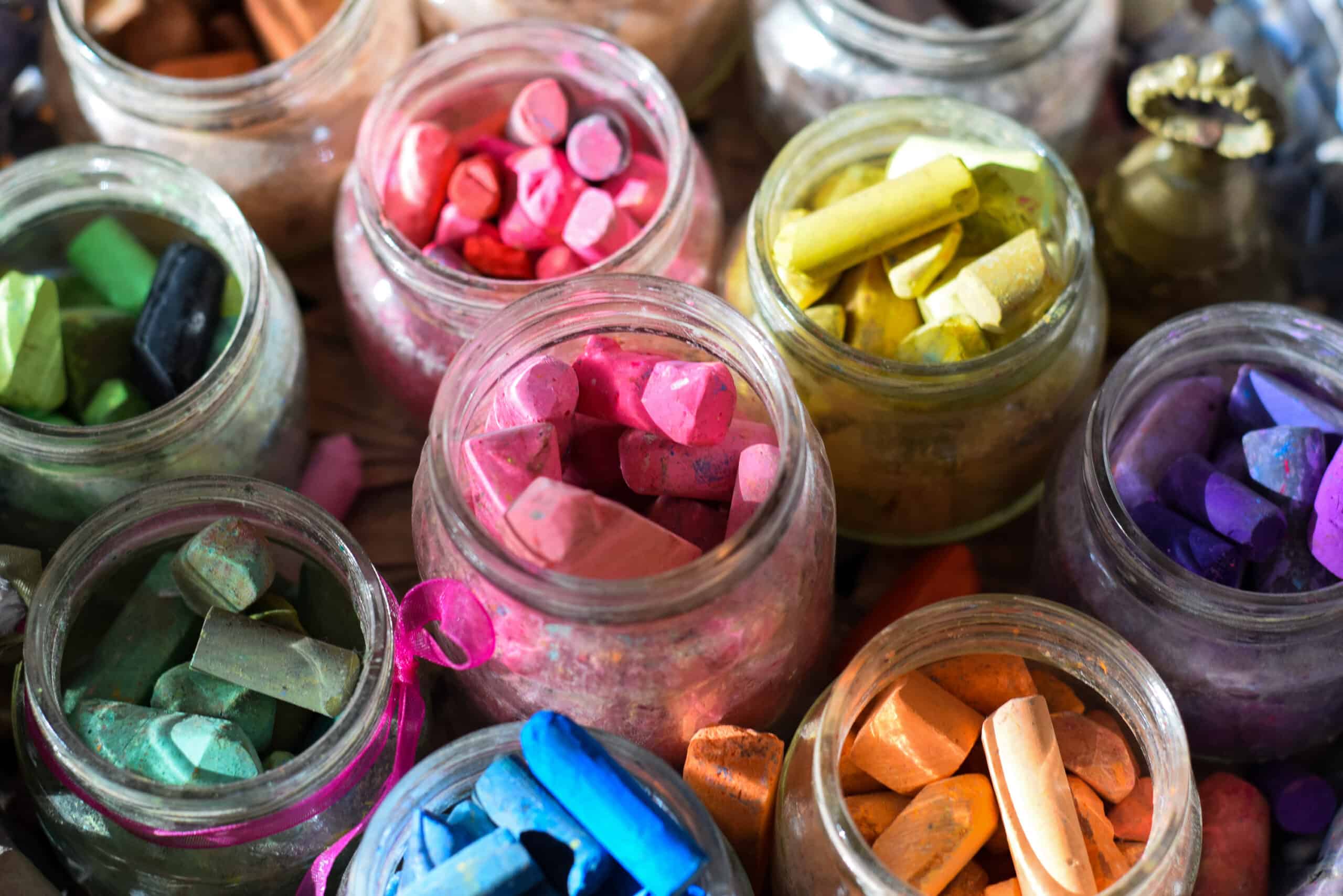 Ten clear glass jars filled with thick pieces of chalk from the top of the frame at 12 o’clock is red or pink and around the circle yellow, purple, orange, blue, light green, and dark green. In the middle of the circle is a red pink color. There are several pieces of chalk in each jar. The jar of light green chalk at 7o’clock has a bright pink ribbon tied around it.