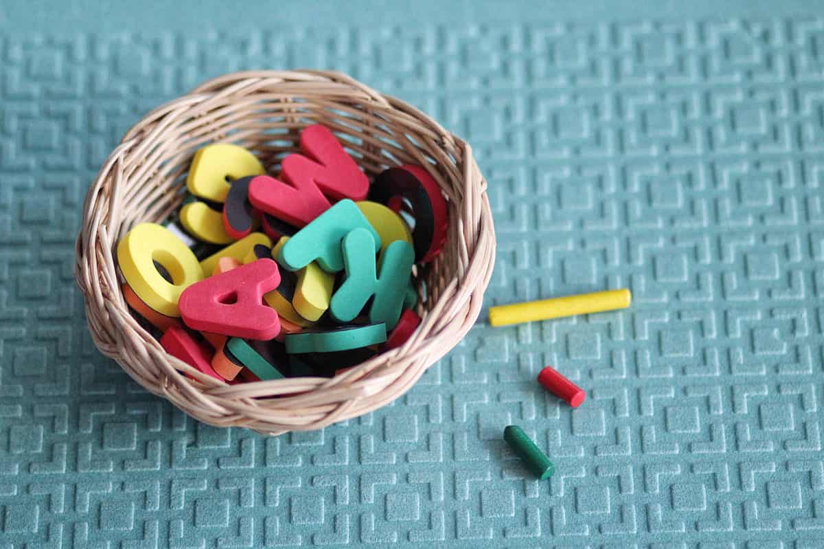 In the left-to-center frame a wicker basket containing uppercase alphabet letters that are red yellow and green. Next to the basket in the right center frame are three pieces of chalk yellow red and green. The yellow piece of chalk is long and appears not to have been used, while the red and green pieces are about a quarter of the size of the yellow stick. Background is a textured light green surface.