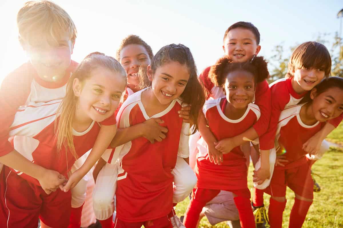 It is very sunny in the left part of the frame.Eight children of various skin tones all dressed in red and white there are four boys  on the backs  of four girls they all seem very happy and are smiling quite beautifully. The boy in frame right has yellow shoelaces on his blue shoes which are the only feet that are visible.