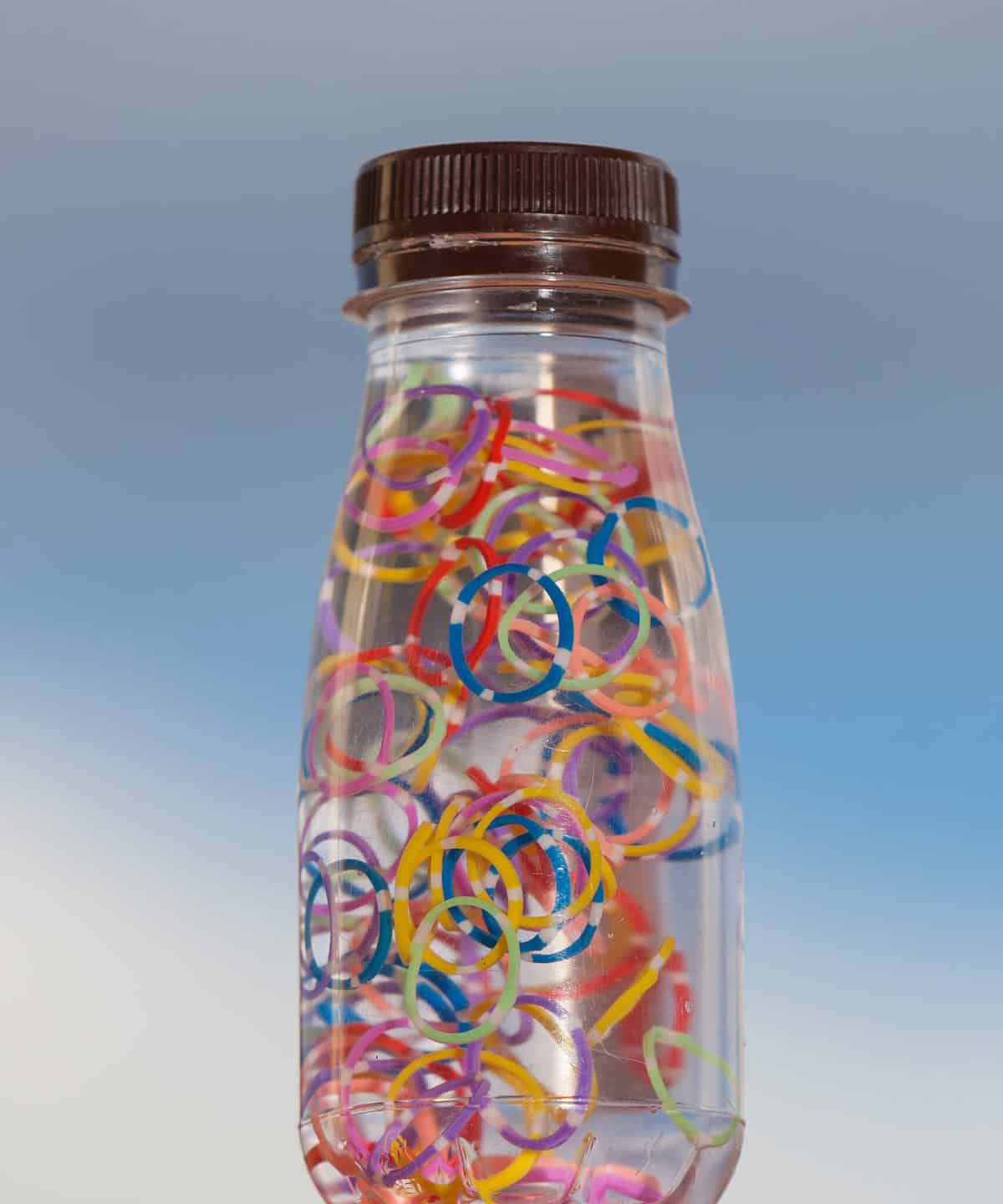 Center frame: a clear glass or plastic bottle with a black plastic top is filled with water and small rubber bands. The rubber bands are green yellow blue red purple and pink. They are all uniform in size. They are small, they type of rubber bands used to create hairstyles with very many braids. The bottle is on a piece of wood. The background is gradient blue.