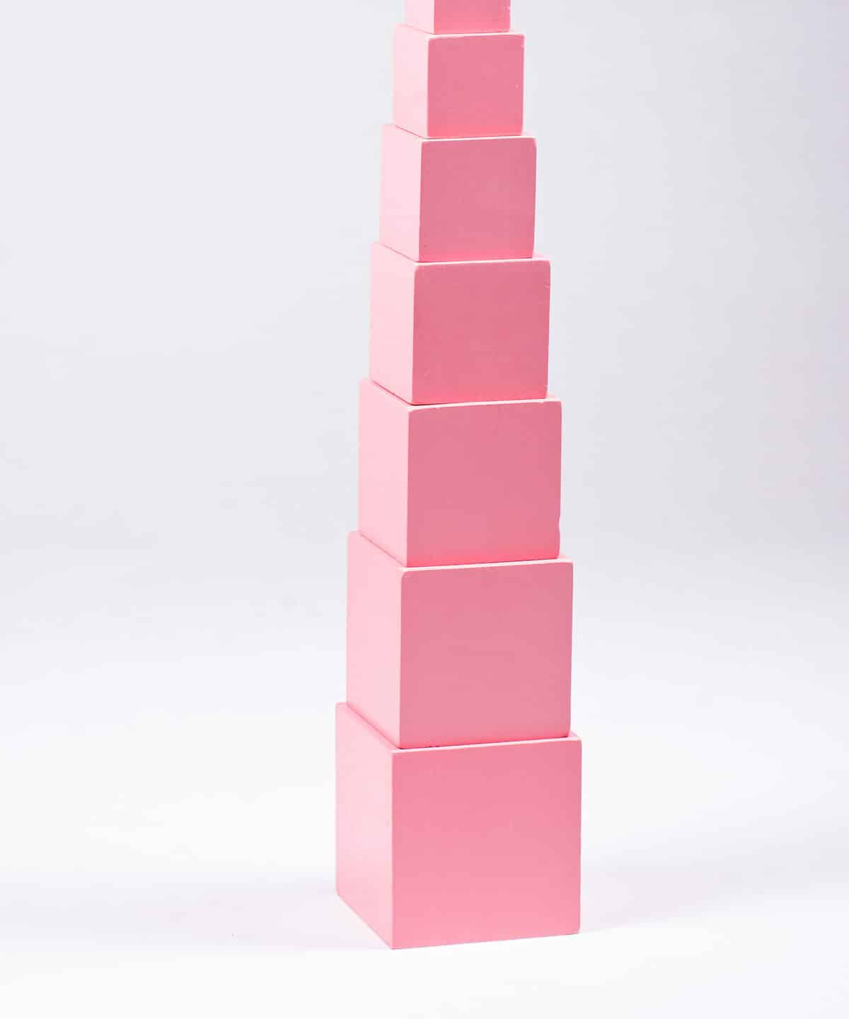 A photograph of the Montessori pink tower. The pink tower consists of 10 graduated blocks that are all painted pink. In the photograph the tower is set up with the largest block on the bottom and the smallest block on the top. It is in the center of the frame. The background is white isolate.