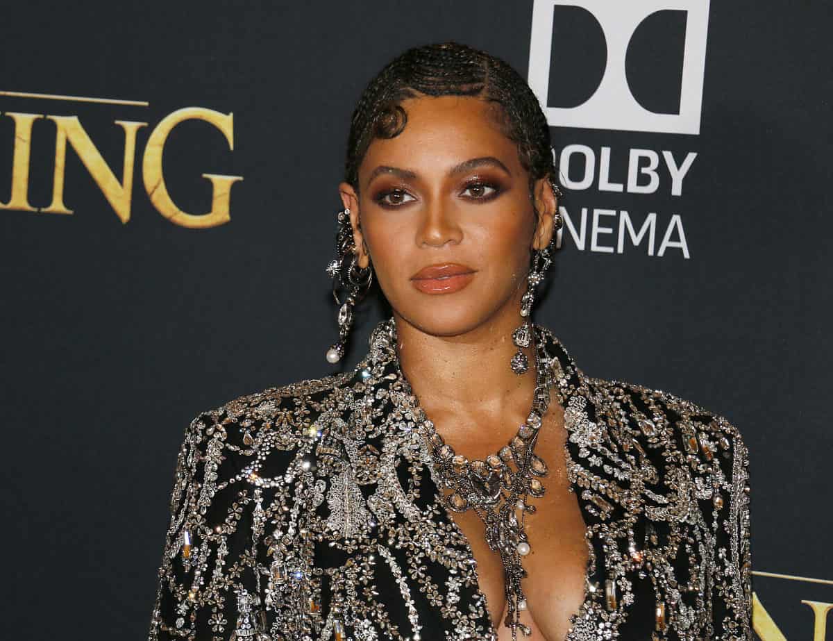 A photograph of Beyonce Knowles in an incredible black jacket with silver thread embroidery and rhinestones. She is also wearing a stunning rhinestone necklace that appears to have some pearls on it it is silver and V-shaped with mini strands hanging off of it that hang in her cleavage. She’s also wearing matching earrings that are approximately 3 inches long. She is light brown skin with black hair that has been styled close to her head. The background is a black screen on which you can see in Golden buffet uppercase letters ING to the left of beyond say space and a little behind her is a sign that says probably Dolby cinema but all you can see is the OLBY Dolby in the NEMA of cinema. And that is white.