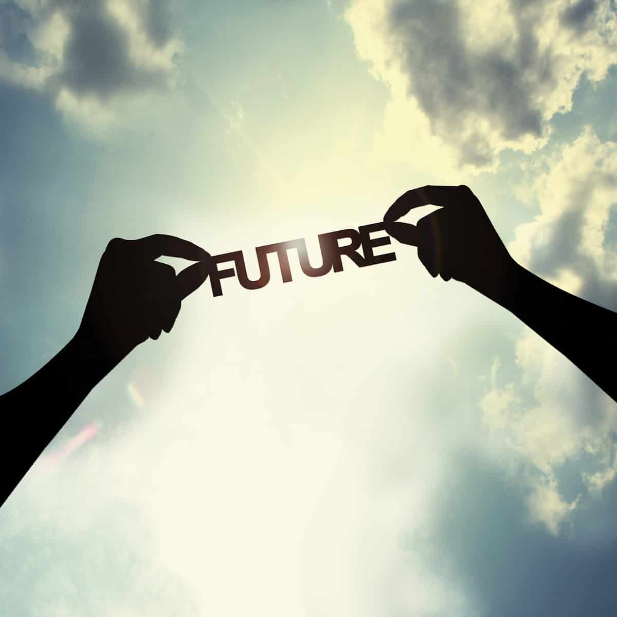 The background of this frame is a bright partly cloudy sky, against which I left arm in the left frame and a right arm in the right frame  in shadow, are visible holding the word future in all uppercase letters that has been cut out of paper or card stock. All of the letters are connected and are about 2 inches tall. 