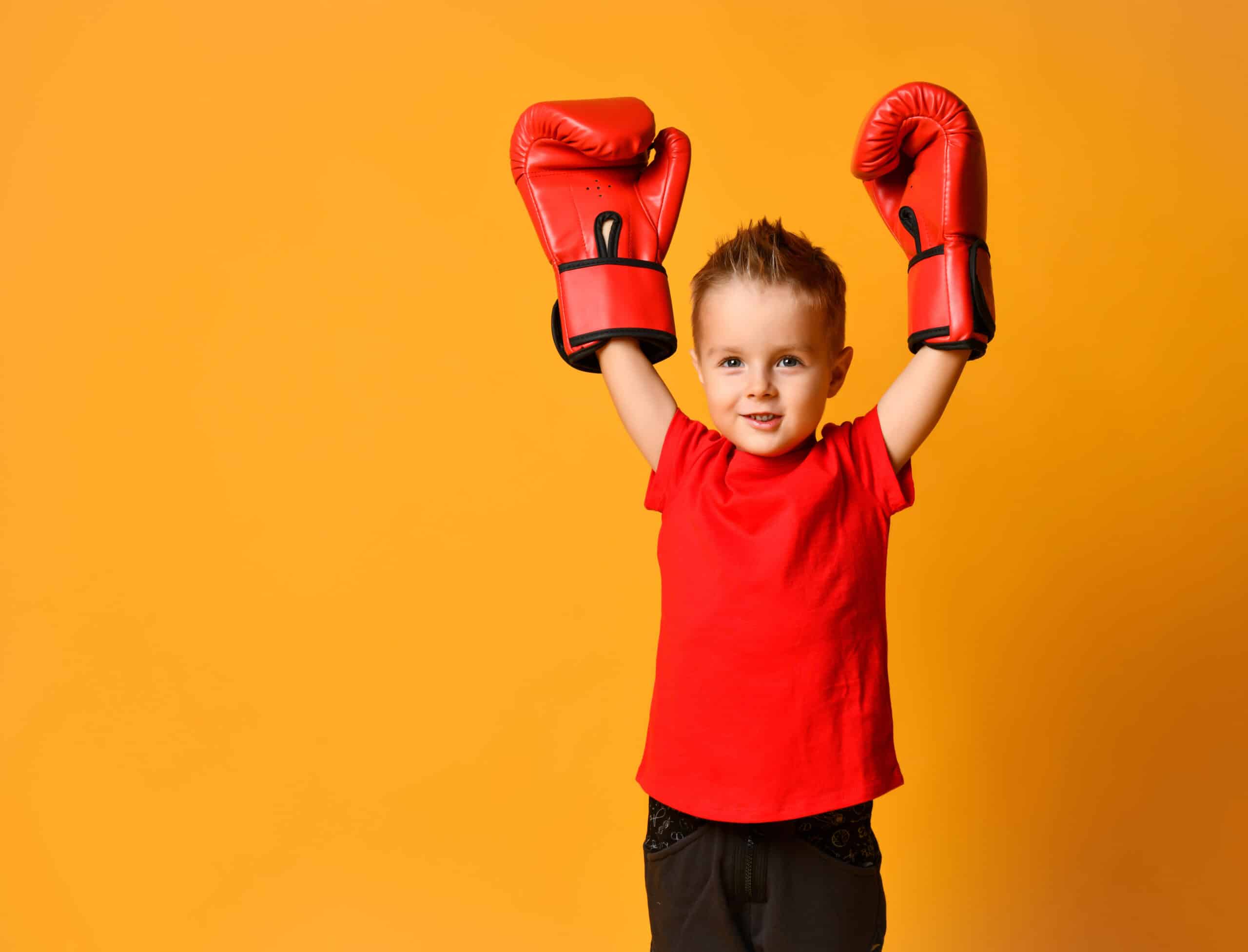 Little Cute Boy 4 years old in a red T-shirt and red boxing gloves on a yellow background, triumphantly raised his hands up, win