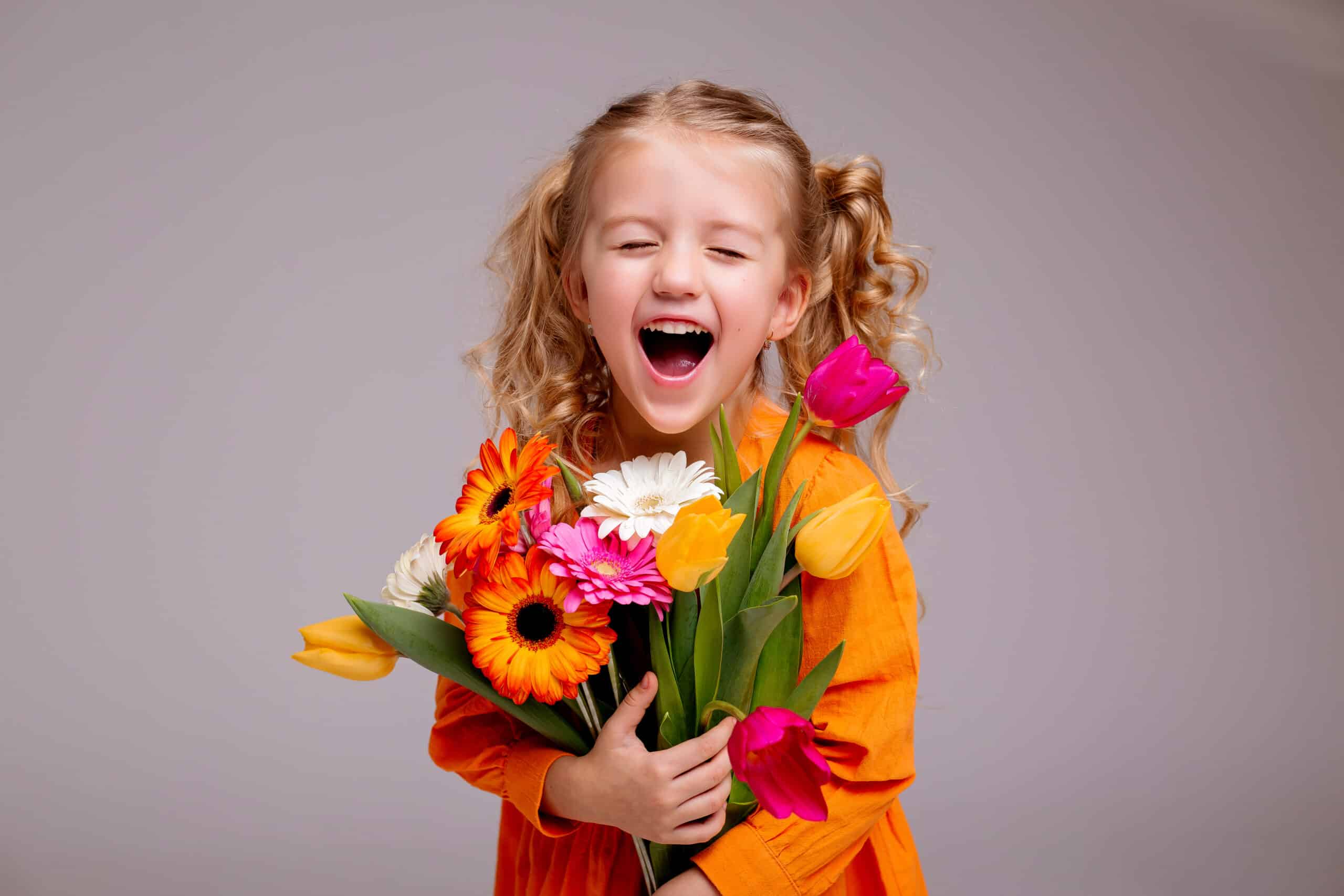 portrait of a little blonde girl with a bouquet of spring flowers on a light background. child in an orange dress holding a bouquet of tulips in his hands. space for text. spring concept
