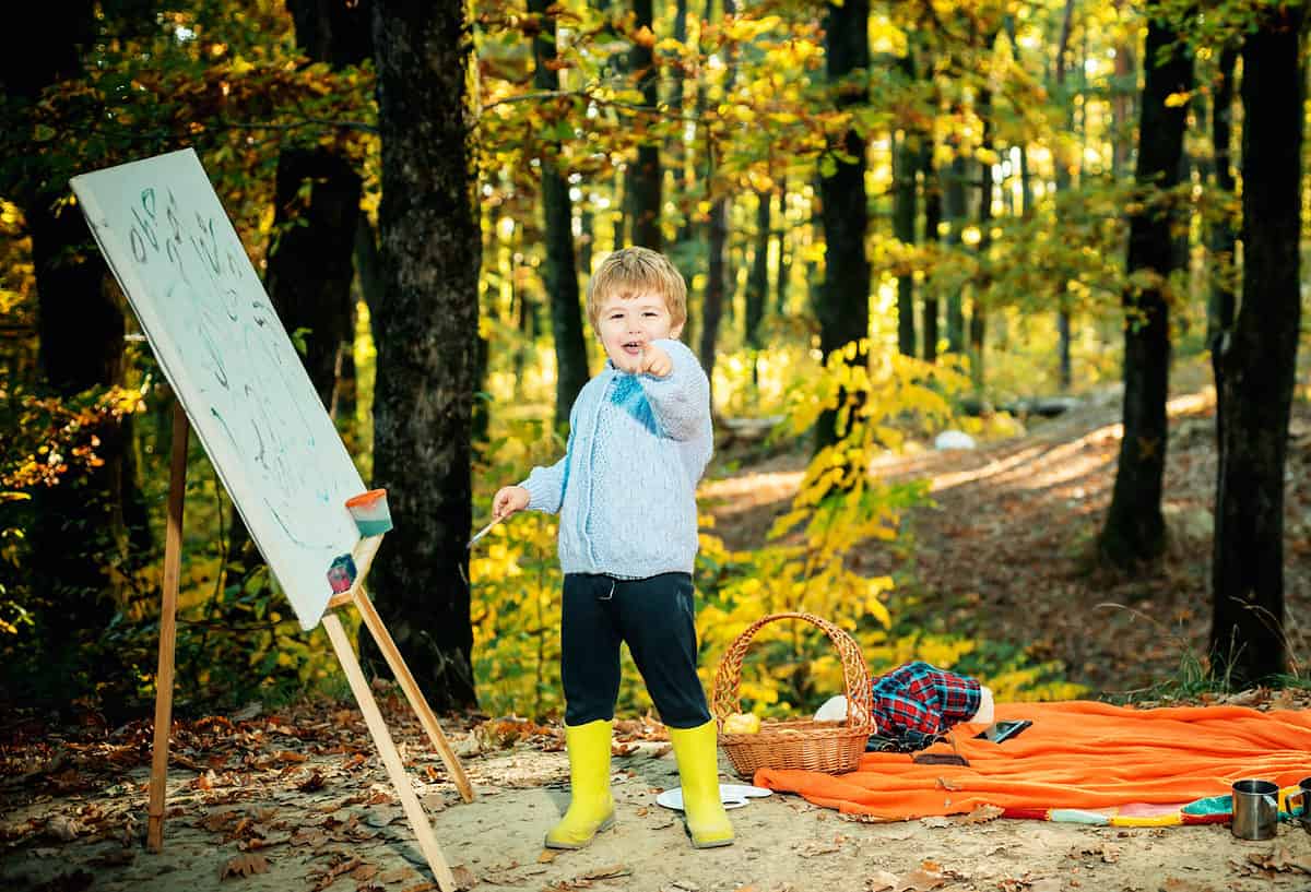 Photo is an outdoor sitting, a wooded area with mini trees in the background with yellow to golden leaves. Center frame is a young child with short strawberry blonde hair wearing a gray button front long sleeve sweater black long pants and yellow rubber boots. They are pointing at the photographer with their left hand while their right hand holds a paintbrush. To the right of the child in the left frame is a wooden easel. On the easel is a large piece of paper on which the child has painted some shapes that are possibly letters in blue or black paint. The majority of the paper remains white. Behind the child on the ground is an orange blanket spread out with a wicker basket and a stuffed animal lying on its face dressed in a plaid robe. There appears to be a smartphone next to the animal.