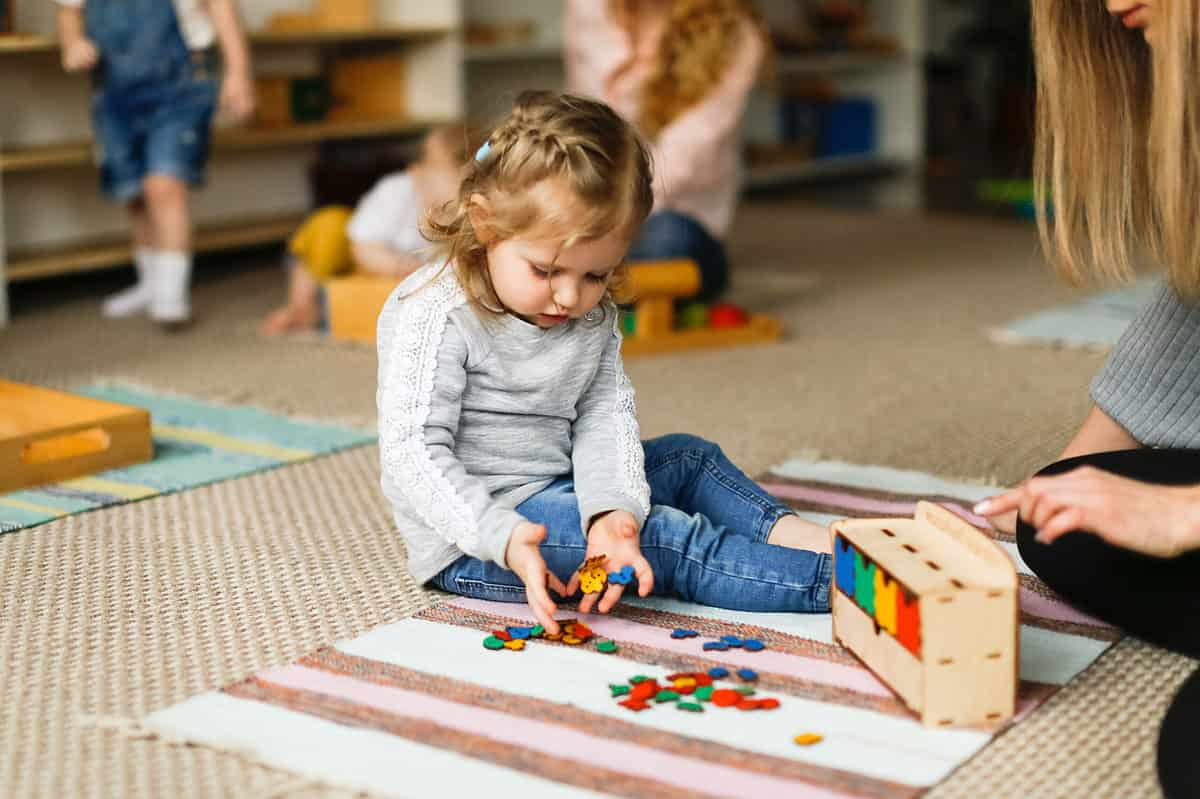 A young girl is seated on a rug center frame the rug is striped with one large white stripe followed by an orangey brown stripe that is not as wide, and then a pink stripe followed by another orangey brown stripe. The pink stripe is a little bit wider than the orangey brown stripes but only about half as wide as the white stripe. The orangey brown stripes are about 1/3 the size of the white stripes. The pattern repeats four times on the small rug. The rug is on carpet that is tan and wheat colored. The young girl is looking down her focus is on her hands which are dropping some colorful manipulatives onto the small rug. They are red yellow blue and green. They are about the size of a quarter, but they are not round. The little girl has on a long sleeve gray shirt with white lace running the length of both sleeves. She is wearing indigo denim jeans. The little girl is the focus of the picture although there are other people in the frame they are out of focus. There are also other rugs visible in the frame. One behind the girl is striped with light blue and green with the light blue stripes being larger than the green ones. A woman with long blonde hair is partially visible on the right side of the frame.