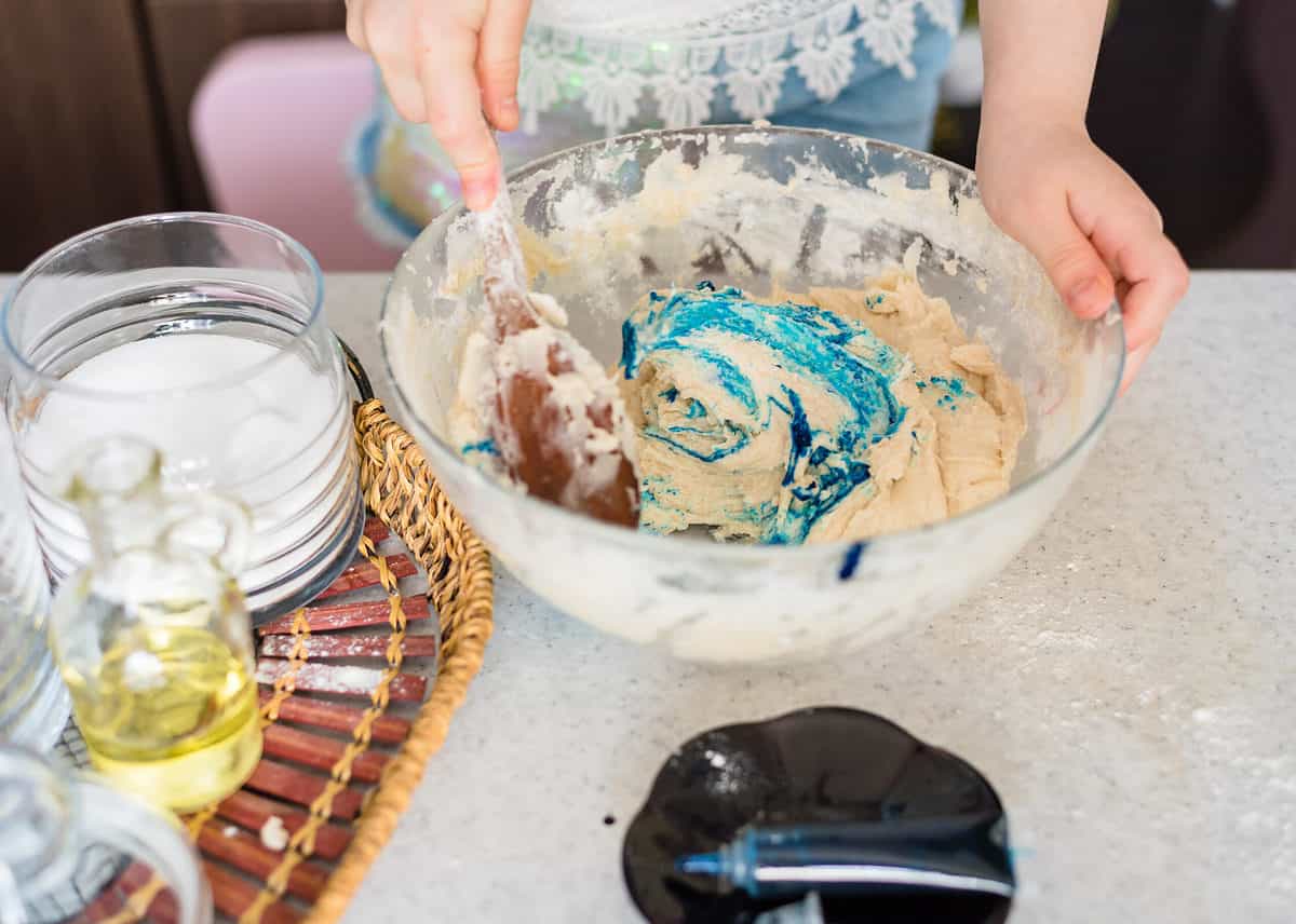 A photograph of a person making play dough. Light skinned hands are visible in the frame. The right hand is holding a wooden spoon. The left hand is holding onto the rim of a glass bowl. The bowl is filled with a doughy looking mixture in which blue food coloring has been squirted but has not been mixed. The tube of blue food coloring is visible in front of the glass bowl it is lying in a black ceramic dish. In the left part of the frame A wooden and woven tray is visible on which flower oil and water are seen.