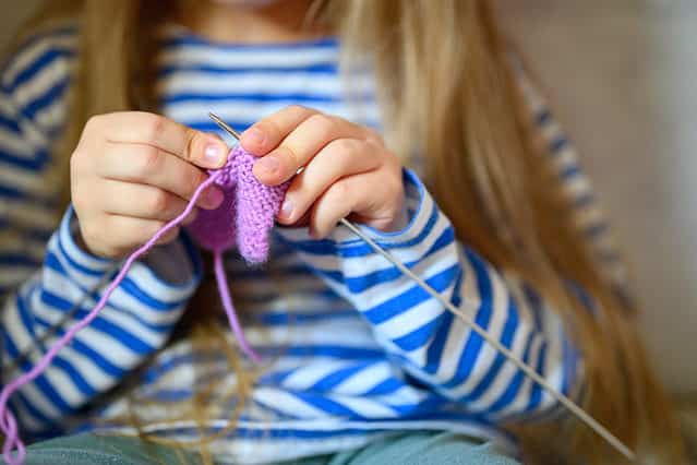 Photograph of a child knitting. All that is visible of the child in the photograph is from the neck to the waist. Child is wearing a long sleeve blue and white striped T-shirt. The child has very long honey blonde hair that falls below their waist. The child is light skinned. The child is knitting with very thin knitting needles using lavender yarn. They have knitted about a 2 inch square which is visible in the Center of the frame between her hands.