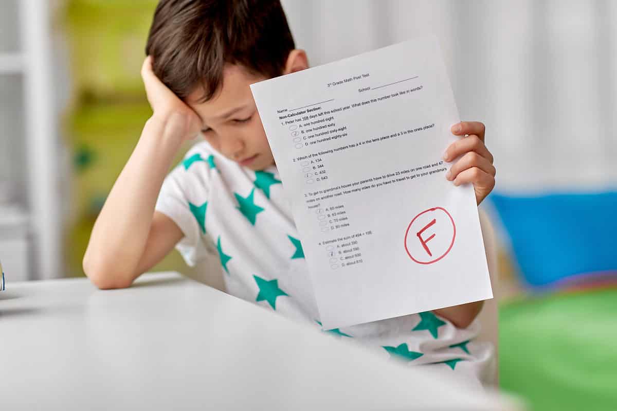 a light skinned male presenting child  looking sad and forlorn,. He is propping up his head with his right hand, though he ihis face is looking down, and his eyes are closed. In his left hand he is holding a  white  sheet of paper with typed multiple choice questions with black letters. On the bottom right side of the test paper is a BIG red uppercase letter F, circled in red. indistinct classroom background with yellow, blue, and green elements. 