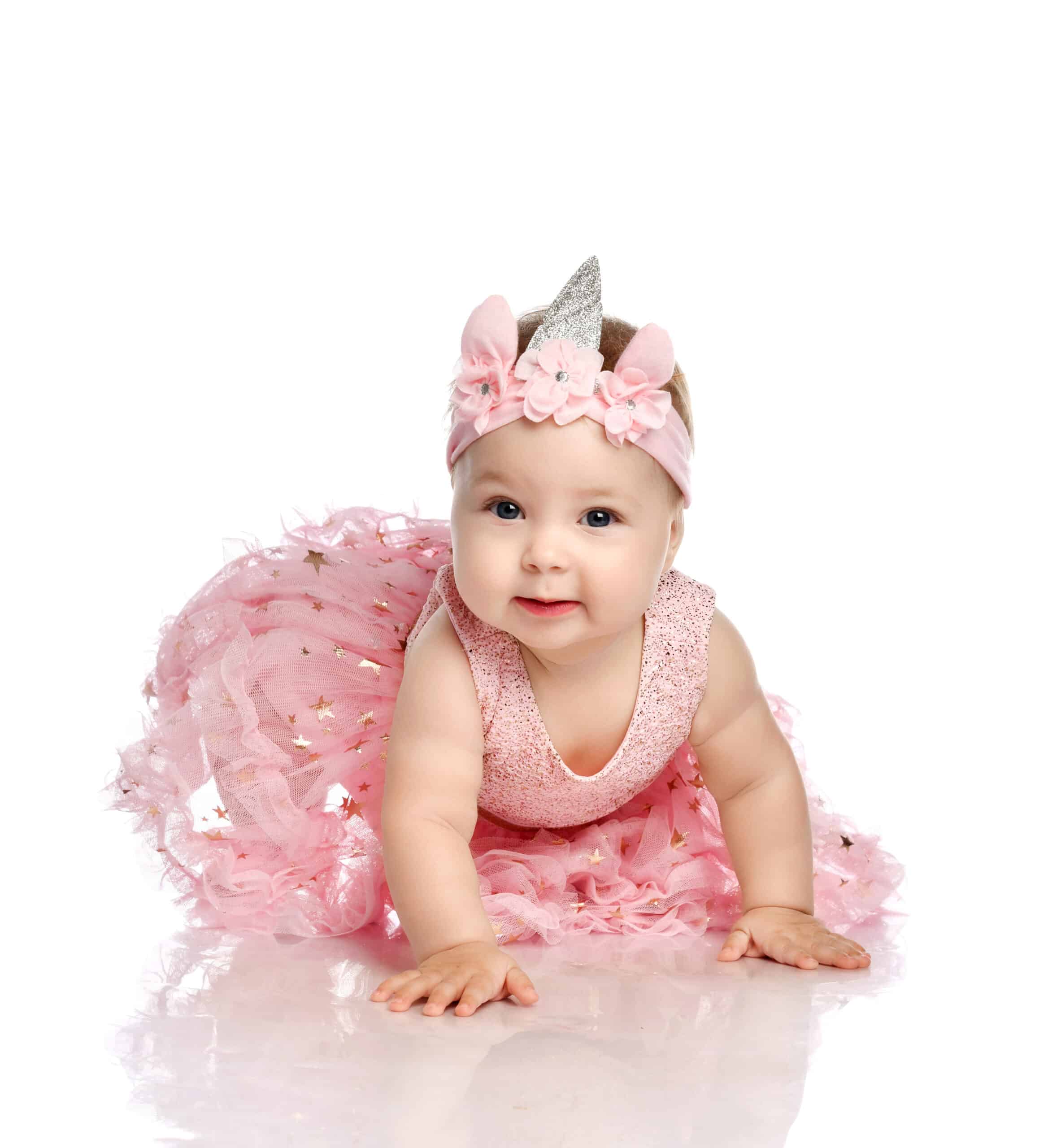 Curious adorable little infant baby girl toddler in a pink dress and crown on head, the costume of the unicorn is creeping on the floor interested in the camera over white background