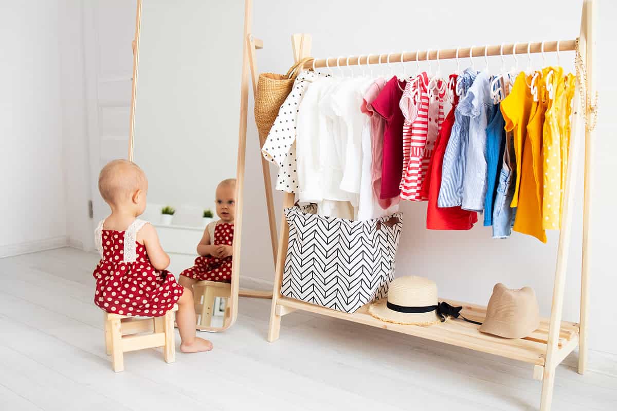 A light-skinned toddler Is visible in the bottom left frame sitting on a natural wooden stool. The toddler is wearing a red sundress with white polkadots. The toddler is facing away from the cameras looking in a full-length natural wood mirror.  The right half of the frame Contains a simple wooden wardrobe that consist of a wooden rod that is about 3 feet long on which several small toddler size clothes are hanging. The clothes are organized in colors from (L-R) white to red to blue to yellow. There is a hamper that is primarily white with a gray chevron pattern on the left side of the wardrobe. Two hats are next to the hamper. The room is completely white including the walls in the floor. Two small green plants in white pots can be seen in the reflection of the mirror.