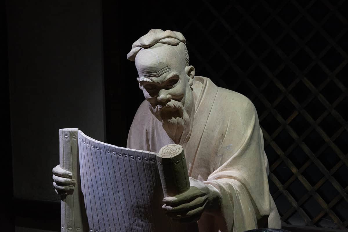 Full frame , black background: A gray stone carving/ statue of Confucius holding a scroll in his hands. His face is tilted down as if he is reading from the scroll. His hair is tied  at the top of his head. He is wearing a flowing robe. 