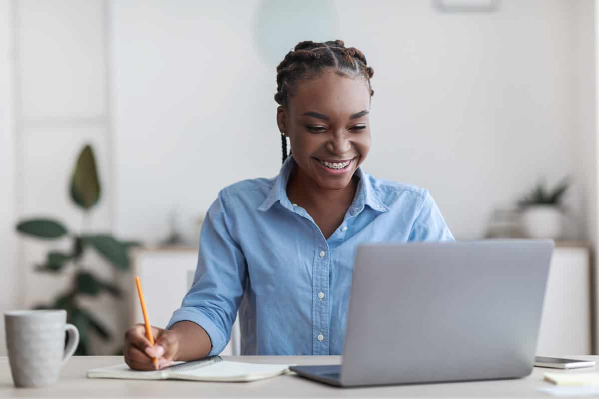 A young brown-skinned woman with cornrows/braids is visible center frame she is wearing a slightly wrinkled light blue oxford cloth button front long sleeve shirt. In her right hand which is in the left frame she is holding a yellow pencil as if riding in a spiral-bound notebook. There is a white coffee mug in the very left part of the frame. In the right bottom part of the frame is a silver laptop computer that is open. The young woman is looking at something on the computer. She is smiling. Her smile reveals that she has orthodontic braces on her teeth. The background is out of focus but is most white with a couple of plants.