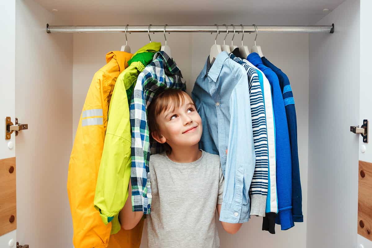 A light skinned male presenting child is seen center frame. He’s standing in a closet surrounded by from the left a yellow raincoat a green windbreaker and a white black green and blue plaid button front shirt. To his left, frame right, or a light blue oxford cloth button front shirt a blue white and gray striped sweater a Carolina blue shirt with a white stripe a royal blue fleece. He is wearing a short sleeved gray T-shirt. He has dark hair that sweeps across his forehead. He is looking up toward right frame apparently at a blue shirt that makes him very happy.