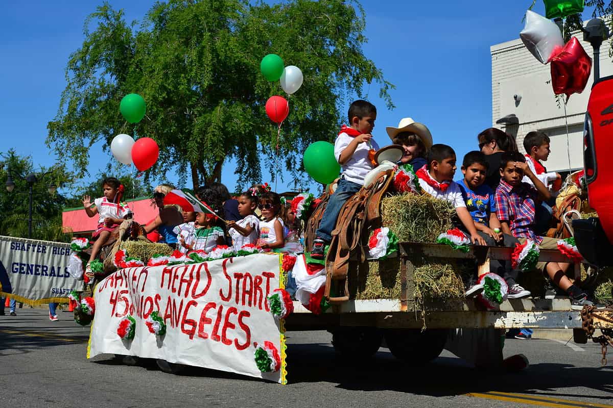 A colorful photograph of a Cinco de Mayo parade float. It has obviously been made by children the who populate the float. A white sign with red letters is visible on the side of the trailer which says Head Start  Los Angeles. There are some other words that are not discernible on the sign. 15 or so brown-skinned and olive-skinned children are riding the float. One adult with olive skin and dark hair wearing a white cowboy hat is also riding the float but is primarily obscured by a little boy wearing a white T-shirt and blue jeans he was sitting on a horse saddle. Many of the children are wearing white shirts and red bandannas however one little boy has on a madras plaid button front shirt and another little boy has on a blue T-shirt with some type of silk screen. The background for the parade float is a beautiful green tree against a vivid blue sky, and the corner of a gray concrete building in the right frame. 