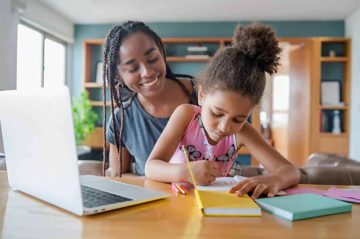 A young brown-skinned girl is seated on her brown-skinned mother's lap. tThe mother is wearing a short-sleeve gray t-shirt, her dark hair is shoulder length braids. The little girl has her hair up in a bun is wearing a sleeveless pink t-shirt. The little girl is holding a pencil in her right hand and appears to be writing on a piece of paper in front of her on a table or desk. There is a yellow book and a seafood green book on the table in from of her. There is a laptop computer visible in the left frame. 