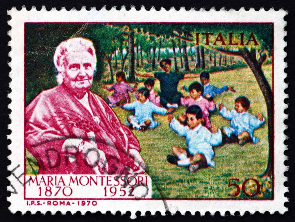 Full Frame: A very colorful Italian postage stamp with white, scalloped edges. An image of Maria Montessori as an elderly woman with white hair, holding a walking stick /cane in her right hand looking toward the frontof the frame, in deep red and white is visible in the left 1/3 of the frame. Behind her, center and right frame are depictions of seven little dark haired children and one dark haired adult seated in green grass, with their arms outstretched at their sides. Trees are visible in the background. It is an illustration, not a photograph. In the upper right frame is the word Italia, in the bottom right corner the number 50, nd in the lower left corner iare the words: Maria Montessori  with the years 1870    1952 below her name. all of the writing is deep red. 