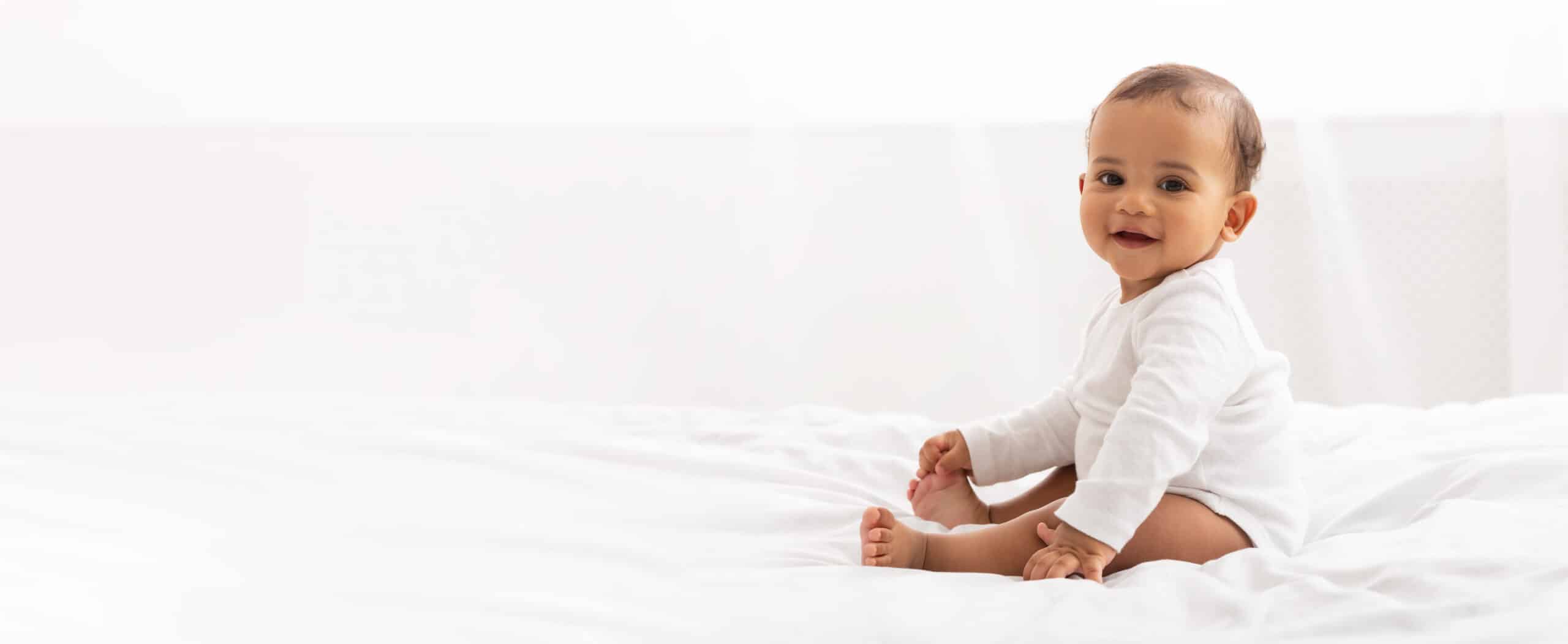 Happy Black Little Baby Girl Sitting On Bed Looking At Camera And Smiling In Bedroom At Home. Advertisement Banner With Cute Child Posing Indoor. Panorama With Copy Space For Your Text