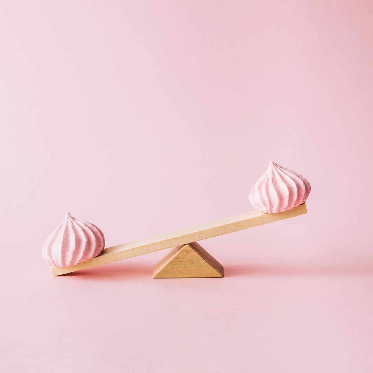 Center frame of a photo is a triangle wedge of wood on top of which is a proportionate size plank of wood. The left side of the wood is down on the pink surface on which the fulcrum is placed. The right side is up in the air. On each end of the liver is a pink macaroon. The whole photo is shot against pink isolate.