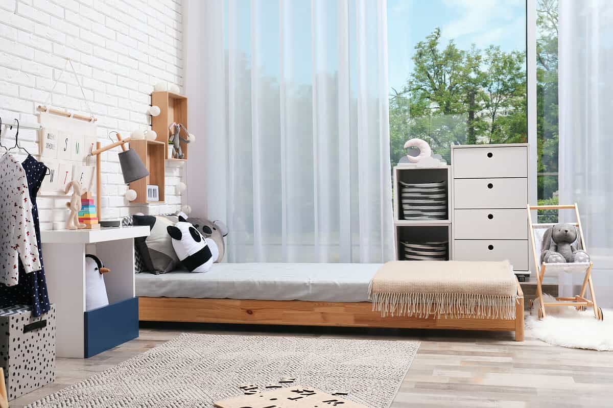 A photograph of a child’s bedroom. On the left side of the frame there are A dress in a shirt visible hanging from a rod near a white brick wall. Next to the clothes hanger is a bedside table that is mostly white with a piece of blue and it’s vase in the front. Above the desk is days of the week hanger with pockets. There is also a wooden lamp with a charcoal gray shade on it. Next to the table is a very low bed made from natural wood on the bed or some pillows and one of the pillows peers to be a panda bear at the end of the bed is a beige blanket with fringe. On the far side of the bed is a wardrobe that is white and consists of two cubes (L) four drawers(R). Next to the wardrobe is a little wooden sling chair with a gray stuffed rabbit sitting in it. There is a natural jute rug on the floor  beside the bed in the front of the frame on with a letter puzzle has been abandoned. The backdrop of the room is a large window with a sheer white curtain that is partially open exposing green trees. 