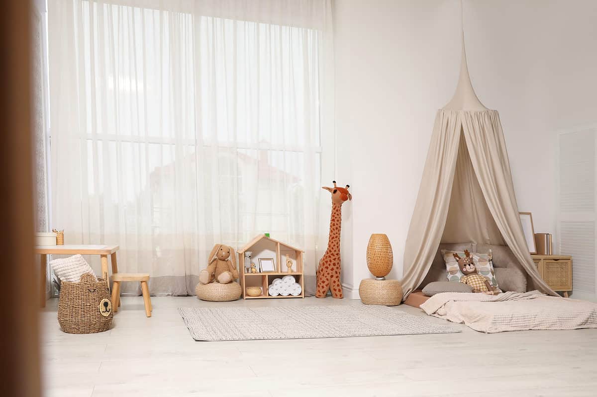 Full frame photograph of a child’s bedroom. Everything in the bedroom has very muted tones from the light gray floor to the white walls. In the left frame is a wicker basket with additions that make it appear to be a bear. There is a wheat colored blanket there’s a bowl at the top. Behind the wicker basket is a child size table with a stool the table and stool our light would the back left to center of the frame is taken up by a very large window over which is a sheer white curtain. In front of the window is a stuffed rabbit perched on a wicker basket next to the rabbit in the very center of the frame is a small doll house with some indiscernible items in it. Next to the dollhouse is a stuffed giraffe that appears to be about 3 feet tall. The right half of the frame is taken up by a bed on the floor that has a light beige canopy over the head of the bed. The bed itself has three taupe pillows And one pillow that is wheat colored with circles of taupe gold and gray. A stuffed brown rabbit with a gold and checkered body leans against the pillow with the circles. There is a neutral colored blanket over the bed. On the far side of the bed is a wicker stand on which a mirror wrist against the wall and possibly some books.