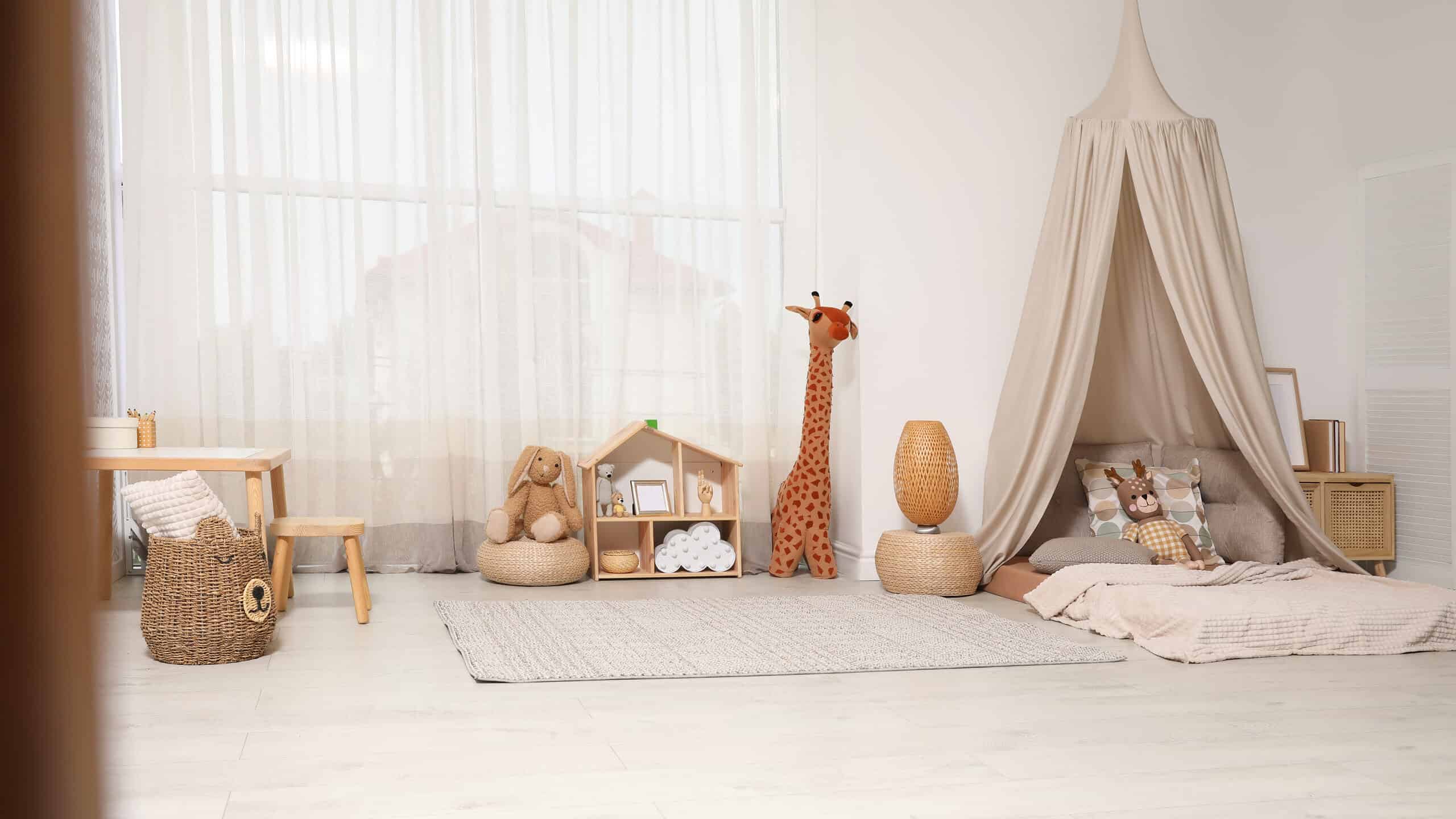 Full frame photograph of a child’s bedroom. Everything in the bedroom has very muted tones from the light gray floor to the white walls. In the left frame is a wicker basket with additions that make it appear to be a bear. There is a wheat colored blanket there’s a bowl at the top. Behind the wicker basket is a child size table with a stool the table and stool our light would the back left to center of the frame is taken up by a very large window over which is a sheer white curtain. In front of the window is a stuffed rabbit perched on a wicker basket next to the rabbit in the very center of the frame is a small doll house with some indiscernible items in it. Next to the dollhouse is a stuffed giraffe that appears to be about 3 feet tall. The right half of the frame is taken up by a bed on the floor that has a light beige canopy over the head of the bed. The bed itself has three taupe pillows And one pillow that is wheat colored with circles of taupe gold and gray. A stuffed brown rabbit with a gold and checkered body leans against the pillow with the circles. There is a neutral colored blanket over the bed. On the far side of the bed is a wicker stand on which a mirror wrist against the wall and possibly some books.