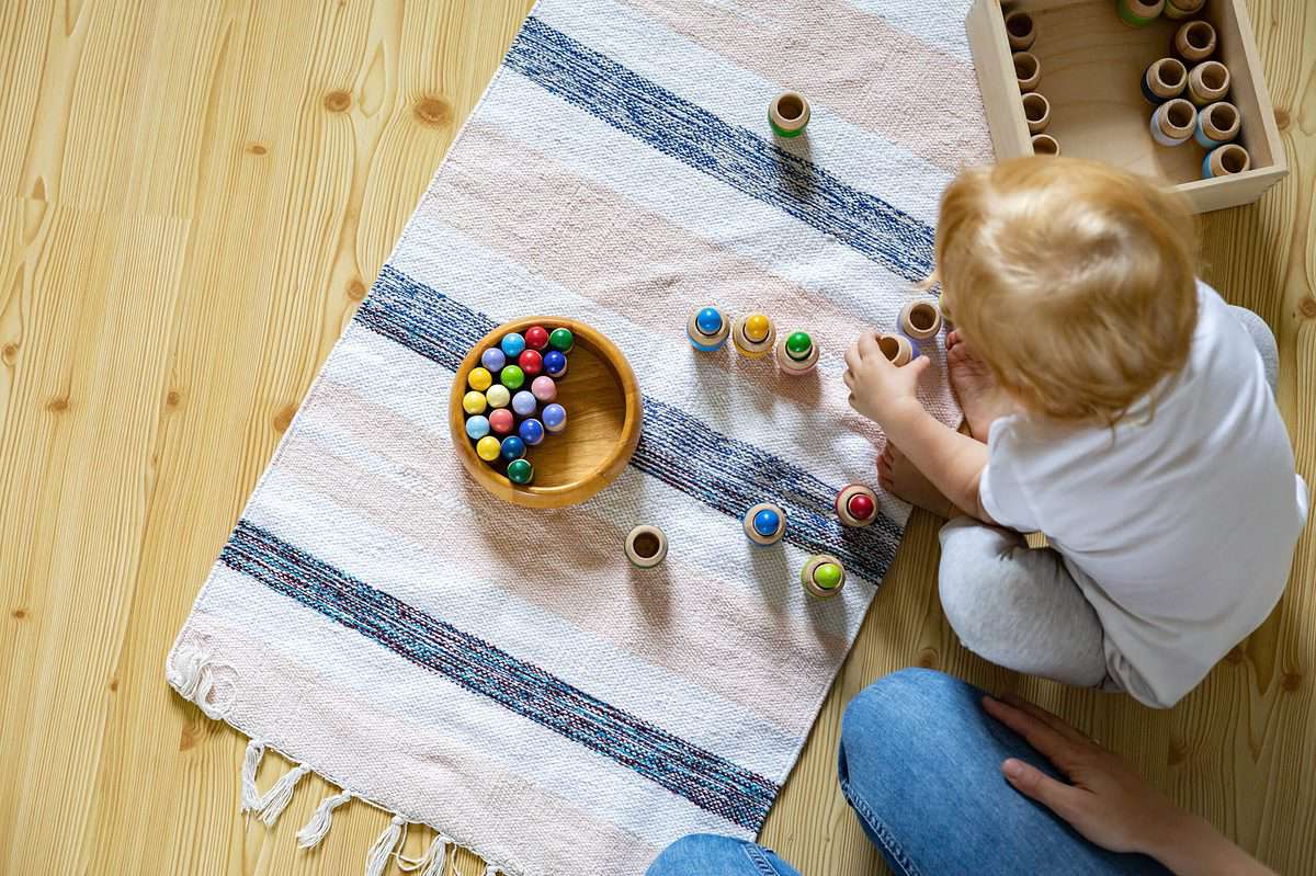 Frame right: a young light skinned child with strawberry blonde hair facing away from the camera is seated on a natural wood floor. In the bottom part of the frame blue Jean clad thighs are visible. A light- skinned hand is resting on the right thigh. In front of the child on the floor is a mat that is striped from a very light flesh color to  white to blue. The pattern repeats four times. On the mat is a natural round wooden bowl with marbles that are yellow, blue, green, and red. There are marble-sizedsmall cups in the mat  in which the young child is placing one marble each. There are 9 cups on the mat six of which have in marble in them. In the upper right frame there is a wooden box with the small wooden cups in it.