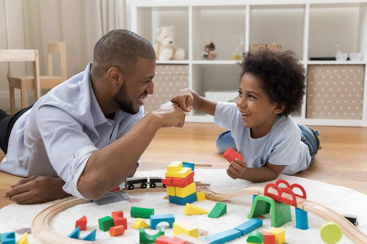 Frame Left: A brown-skinned man with very close cropped hair and a short beard wearing a button front light blue oxford cloth shirt and black pants is lying on the floor which is wooden one arm on a white carpet that also has a round wooden train track, is visible, fist bumping A young brown skin boy who is in frame right. The boy is wearing a white short sleeved T-shirt and light blue denim jeans. His hair is unruly curls. In his left hand he is holding a rectangular red wooden block. Both he and the man are smiling as if happy. The background consists of white shelves on which there is a teddy bear and a little doll there is also a wooden table and chair and some white curtains