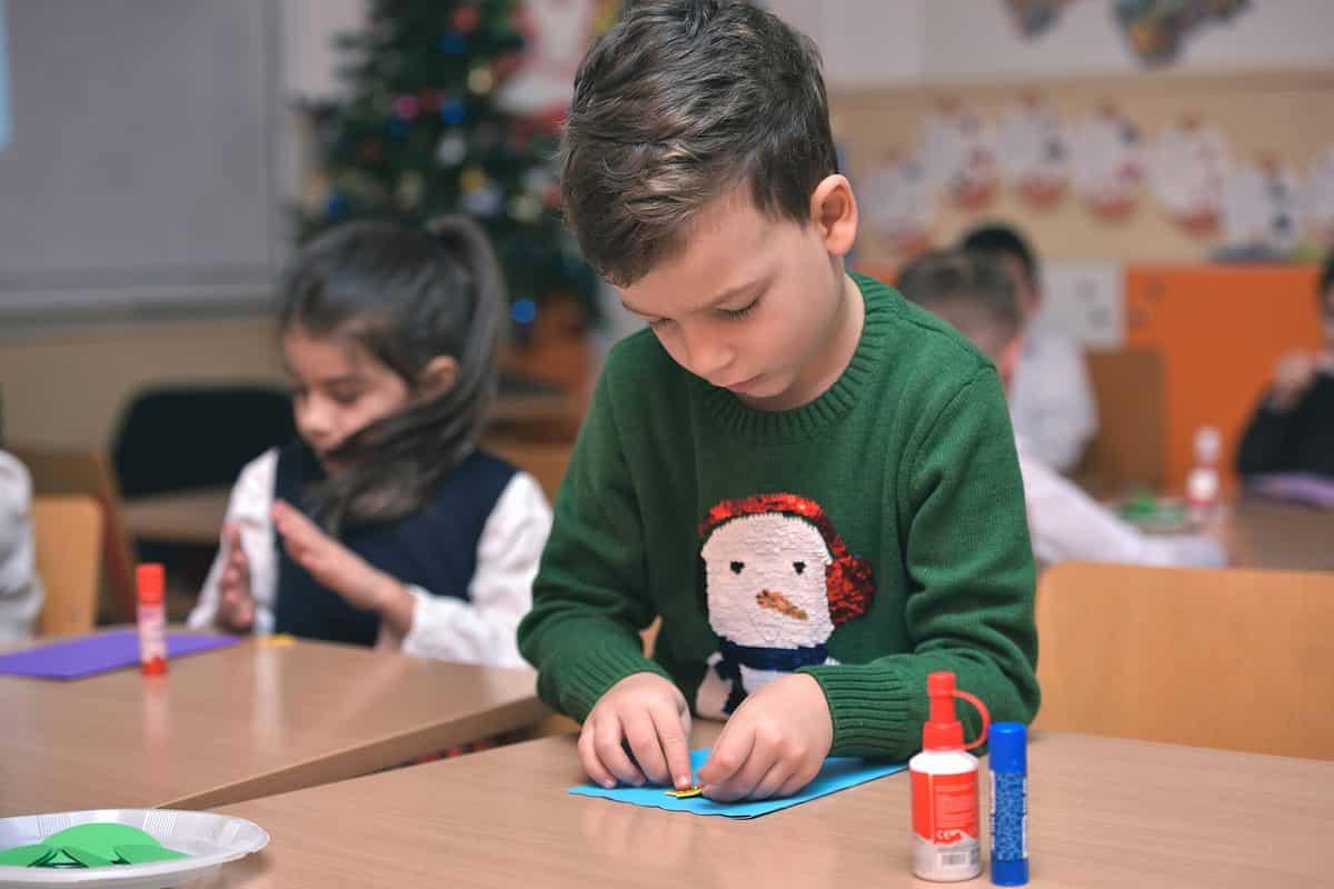 A photograph in a classroom setting at Christmas time. Center frame a young boy is visible concentrating on gluing a yellow piece of paper onto a blue piece of paper. He is light skinned with short brown hair. He is wearing a forest green sweater with the image of a snowman on it. The snowman is white with an orange carrot nose to black coal eyes and a pair of red earmuffs. Behind him is a little girl with long dark hair and light skin wearing a long sleeve white blouse and a black sleeveless sweater. She too is concentrating on gluing paper. The background is an out of focus classroom with a Christmas tree in the corner. 