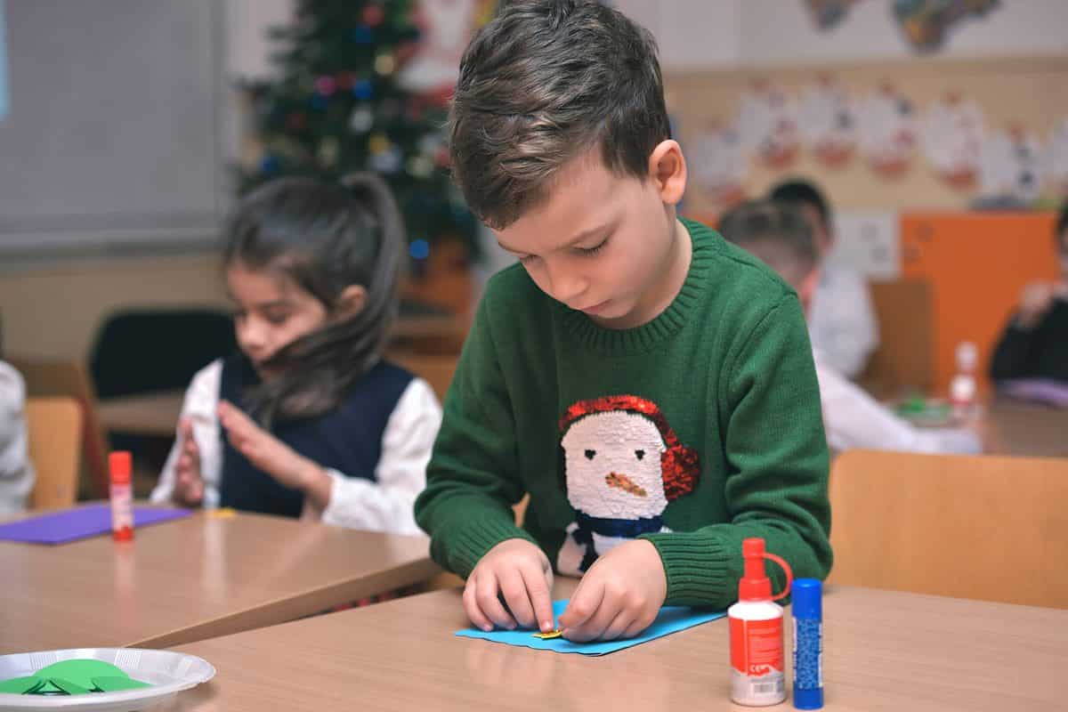 A photograph in a classroom setting at Christmas time. Center frame a young boy is visible concentrating on gluing a yellow piece of paper onto a blue piece of paper. He is light skinned with short brown hair. He is wearing a forest green sweater with the image of a snowman on it. The snowman is white with an orange carrot nose to black coal eyes and a pair of red earmuffs. Behind him is a little girl with long dark hair and light skin wearing a long sleeve white blouse and a black sleeveless sweater. She too is concentrating on gluing paper. The background is an out of focus classroom with a Christmas tree in the corner.