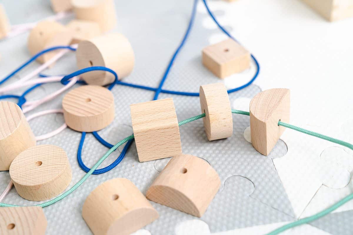 Full frame of natural colored wooden beads. Some of the beads are round some of them are square and some of them are semi circle shaped. The frame consists of about 15 beads of which three are strong on a seafoam green cord. There is also blue cord and pink cord visible in the frame but they do not have anything strung on them. The background consists of cardboard interlocking pieces that are white and gold checked.